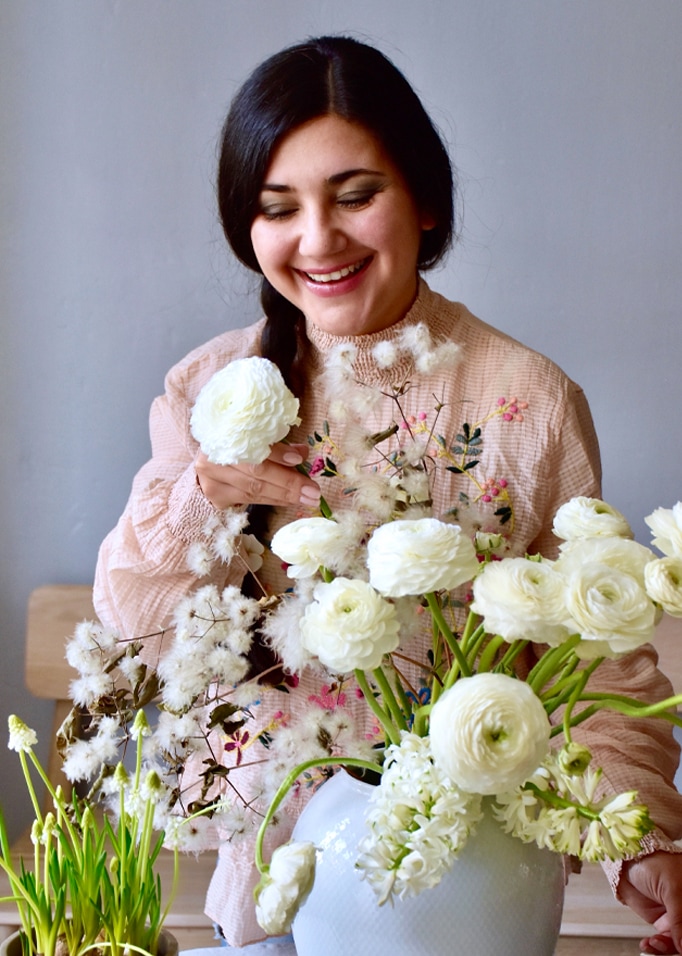 Frau in pastellfarbener Bluse arrangiert weiße Blumen in Vase vor hellem Hintergrund