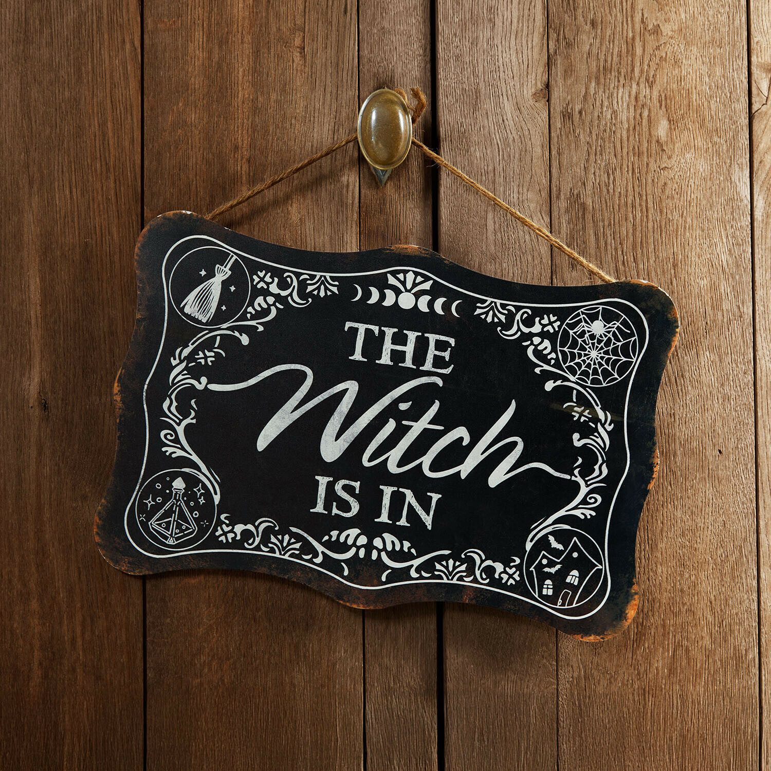 Deko-Schild Talmora Schwarzes Holzschild mit Aufschrift The Witch is in und weißen Verzierungen vor Holzwand