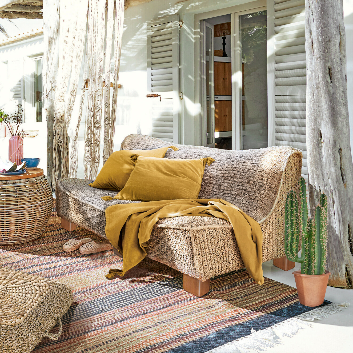 Sofa Bocage Sofa aus geflochtenem Material in Naturfarbe mit senffarbenen Kissen und Decke auf sonniger Terrasse