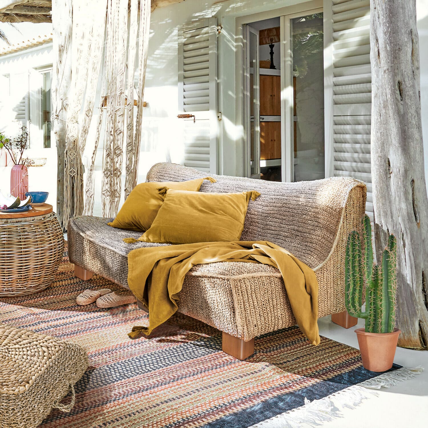 Sofa Bocage Sofa aus geflochtenem Material in Naturfarbe mit senffarbenen Kissen und Decke auf sonniger Terrasse