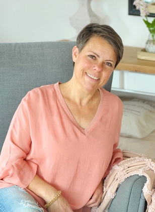 Lächelnde Frau in rosa Bluse sitzt auf grauem Sofa in hellem Raum