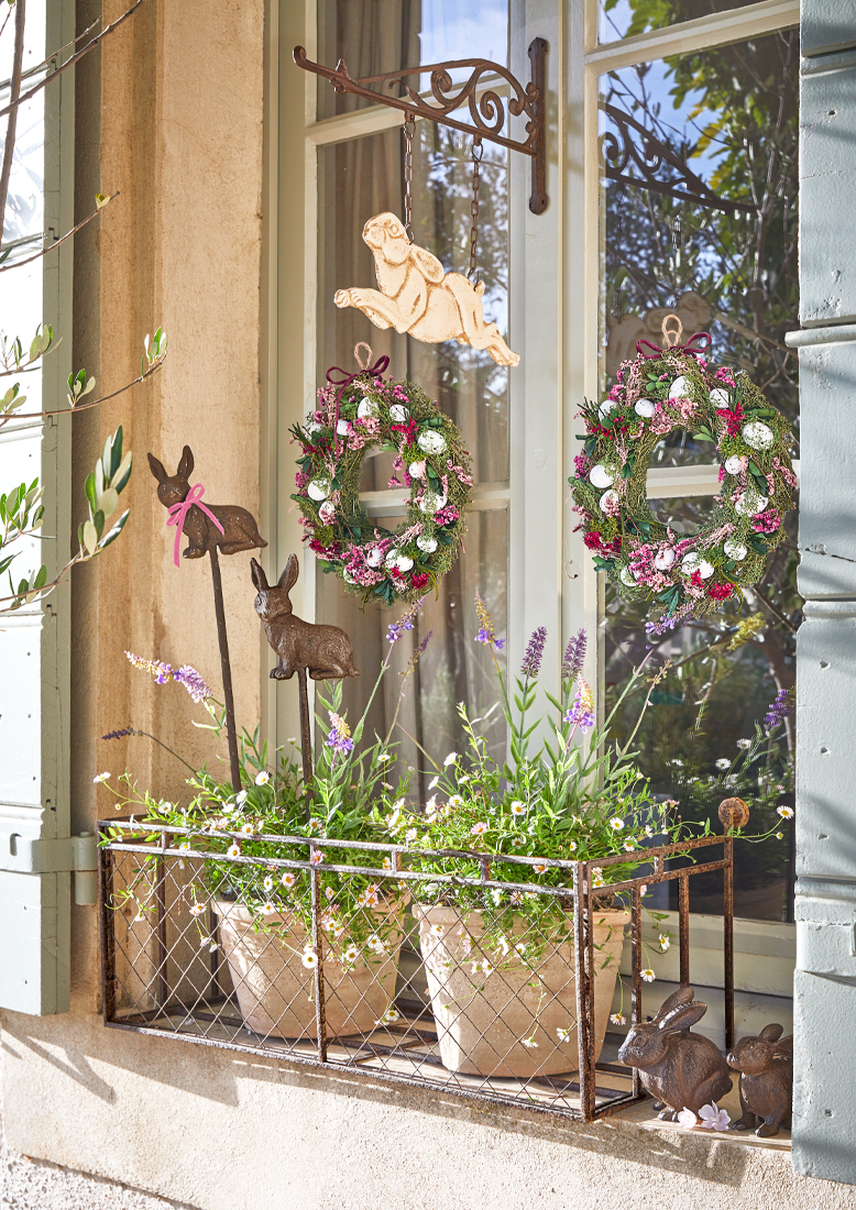 Fensterdekoration mit Metallhalter, Blumentöpfen und Frühlingskränzen mit Eiern und Hasenfiguren