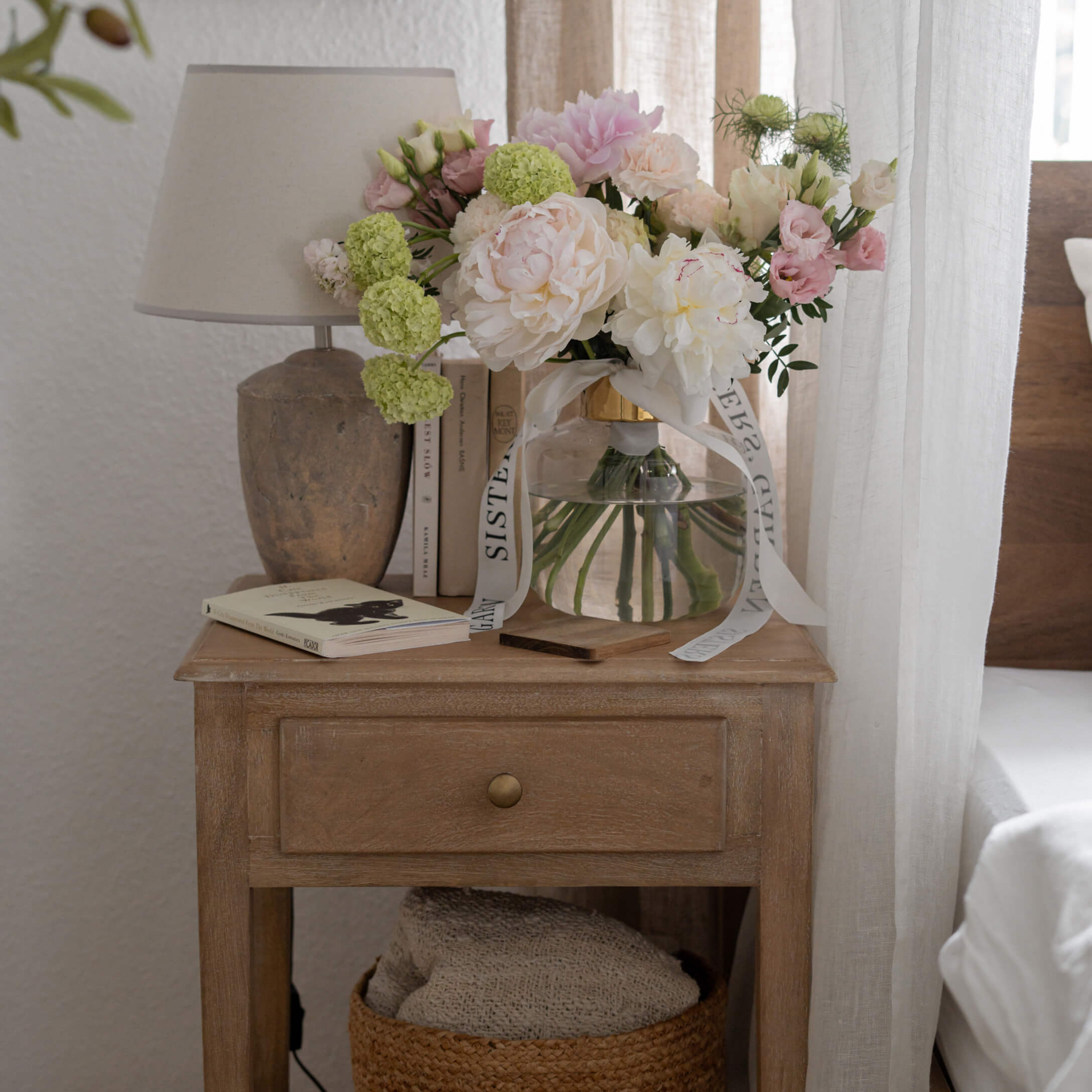 Holz-Nachttisch mit Schublade, Lampe und Vase mit pastellfarbenen Blumen in hellem Raum