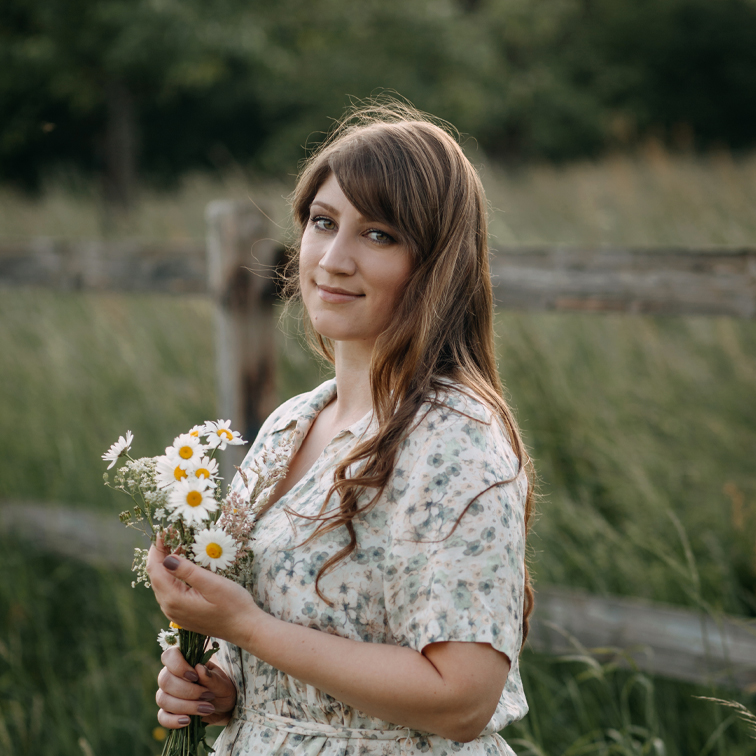 Frau in geblümtem Kleid hält einen Strauß Gänseblümchen vor grünem Feld und Holzzaun