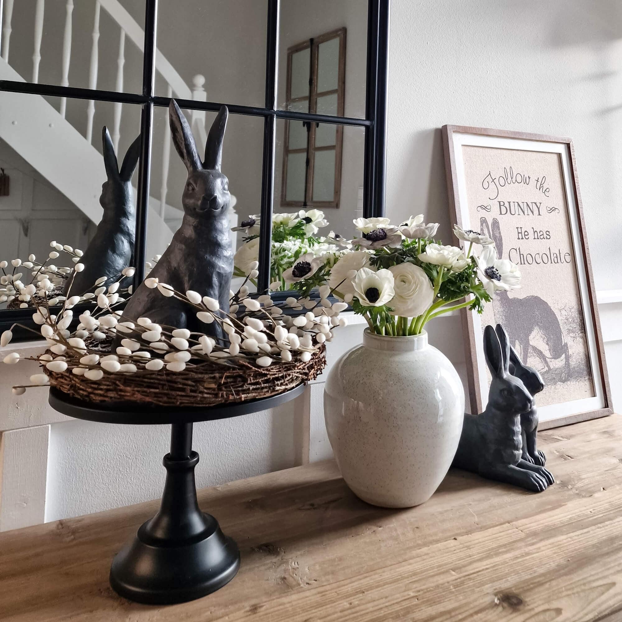 Dekorativer schwarzer Hase mit Weidenkranz und weißen Blumen in Keramikvase auf Holztisch