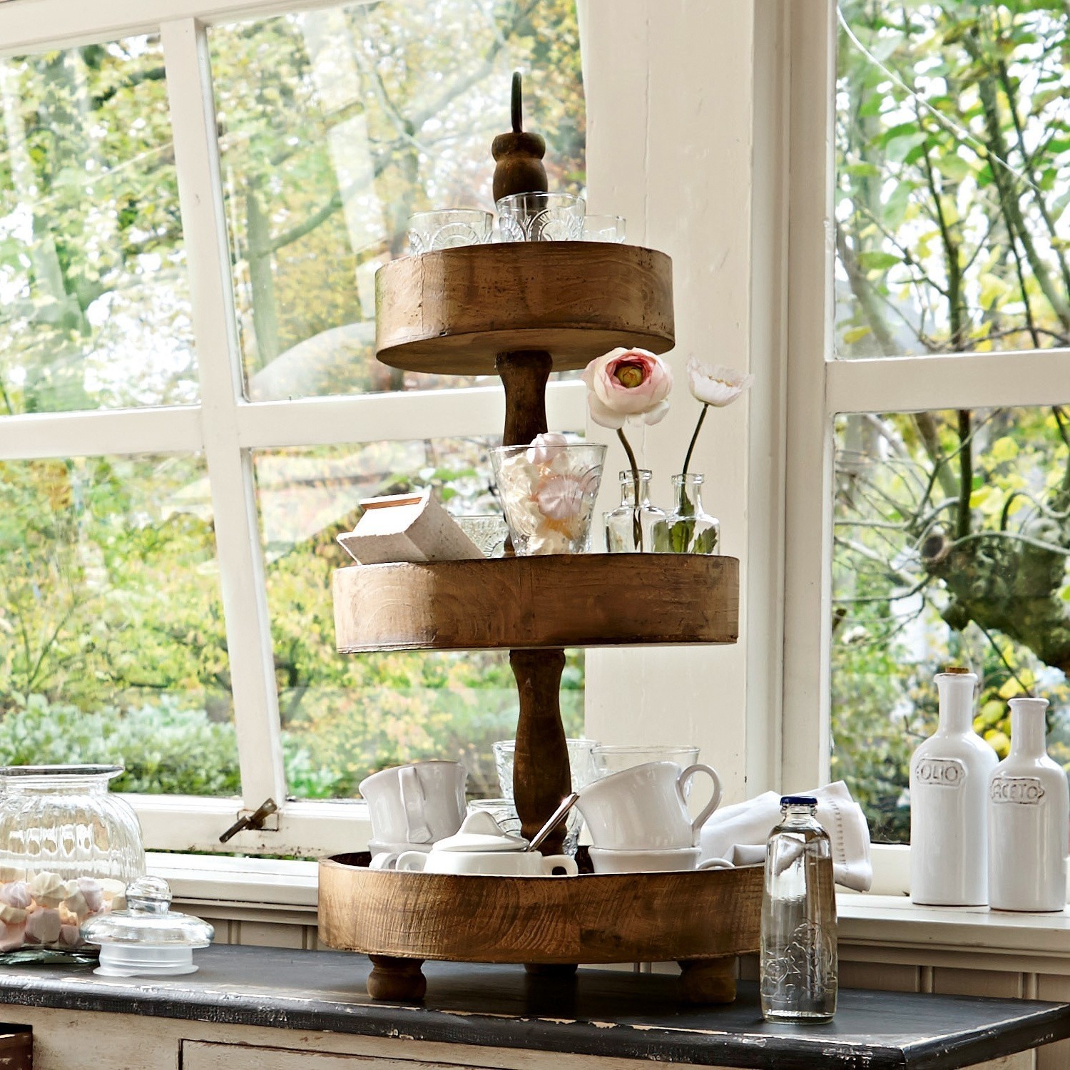 Dreistöckiges Holztablett in Naturfarbe mit Tassen und Dekorationen vor Fenster mit grünem Gartenblick