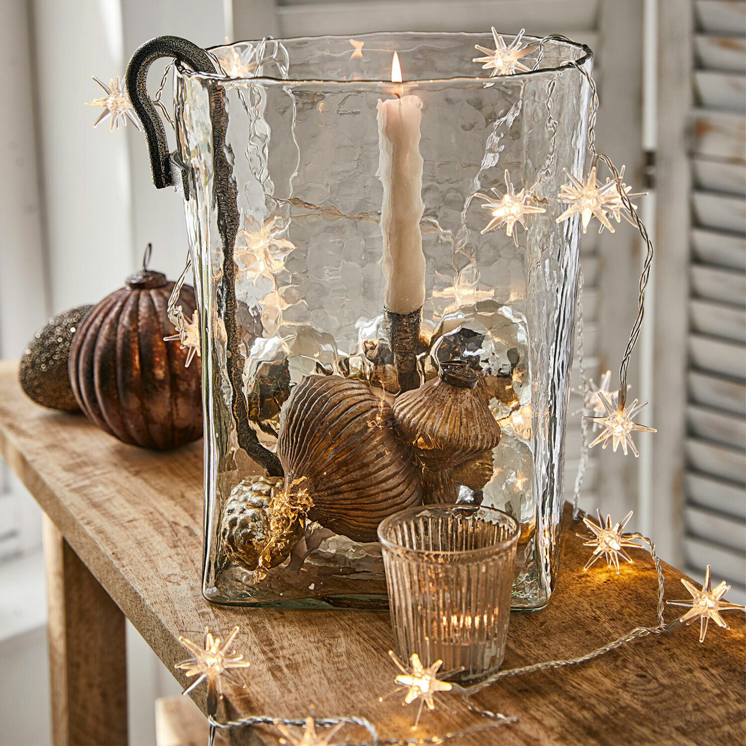 Lichterkette Nelisse Glas-Windlicht mit Metallgriff und kupferfarbenen Dekorationen auf Holztisch mit Stern-Lichterkette