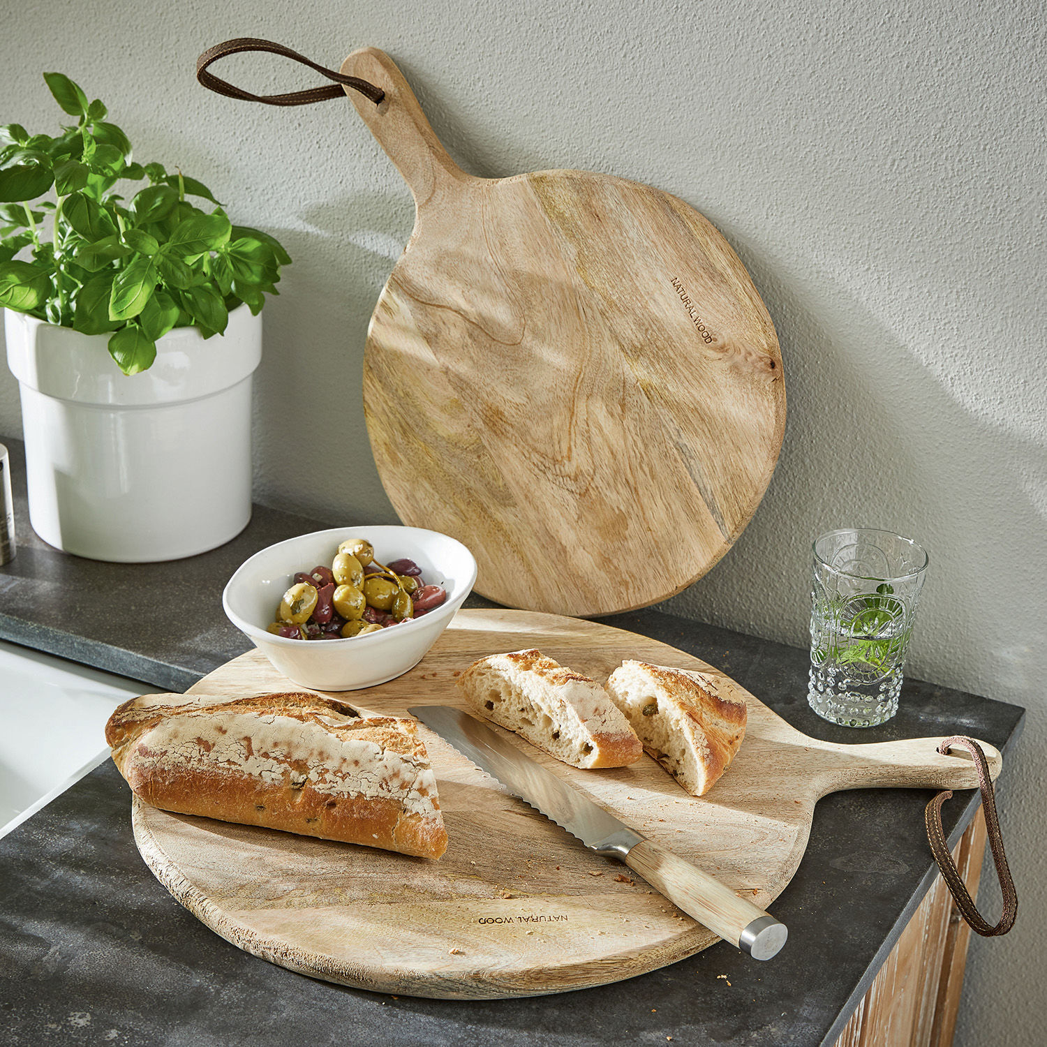 Schneidebrett 2er Set Nitra Runde Schneidebrett aus Naturholz mit Griff, Brotmesser, Brot und Oliven auf Küchenarbeitsplatte