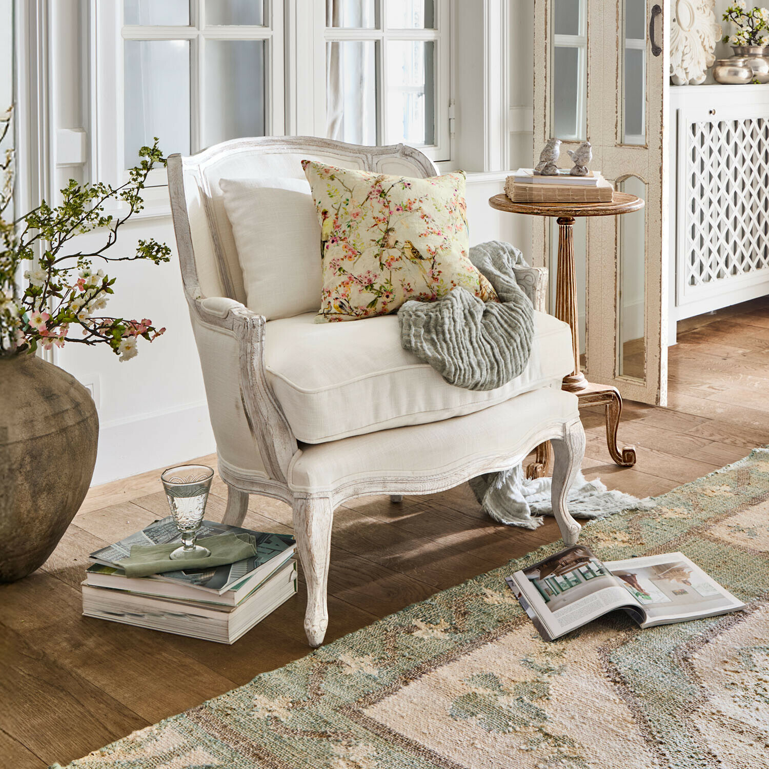 Weißer Holzsessel mit heller Polsterung, floralem Kissen und grauer Decke in gemütlichem Wohnzimmer