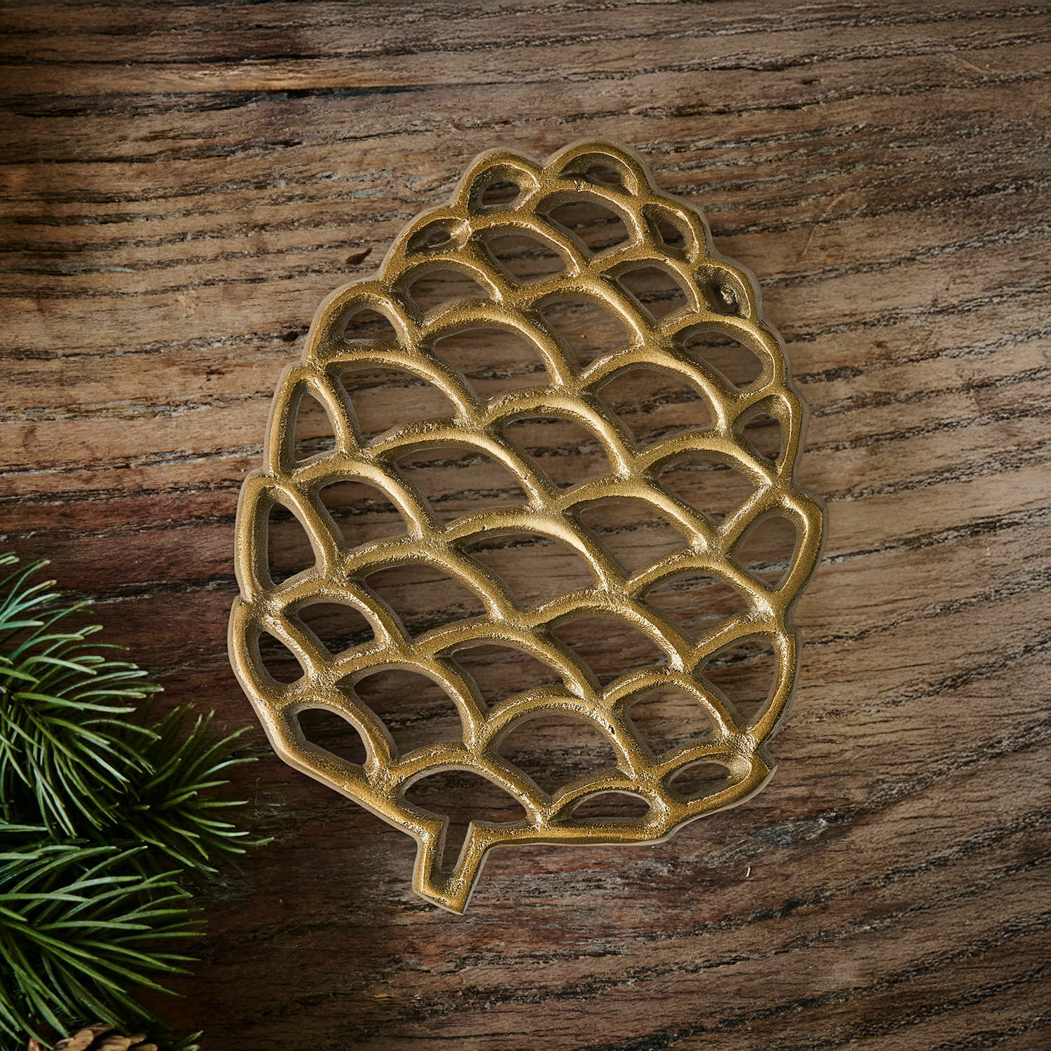 Goldfarbener Metall-Untersetzer in Zapfenform auf Holztisch mit grünen Zweigen
