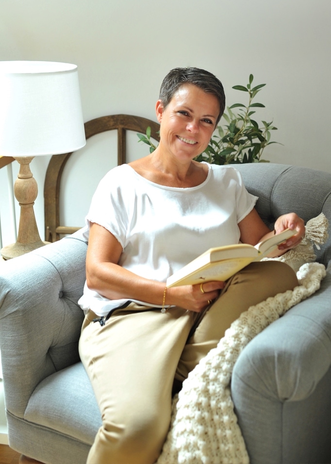 Frau liest Buch auf grauem Polstersessel mit Lampe und Pflanze im Hintergrund