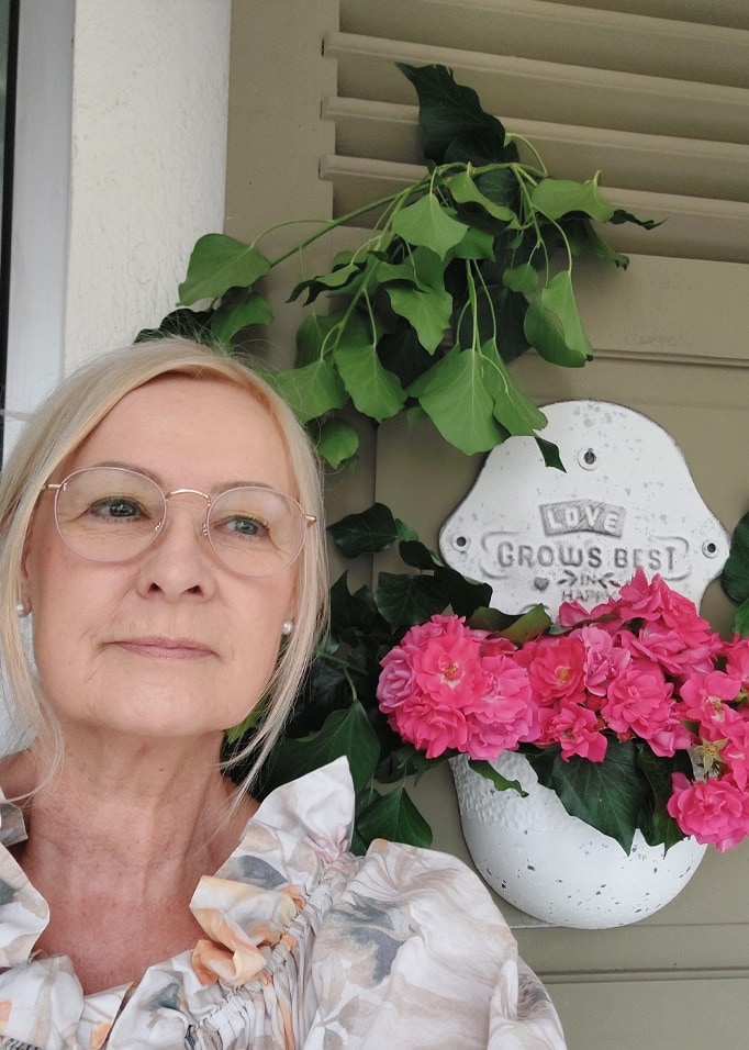 Weiße Wandblumenvase mit pinken Blumen und grünen Blättern vor grauen Jalousien