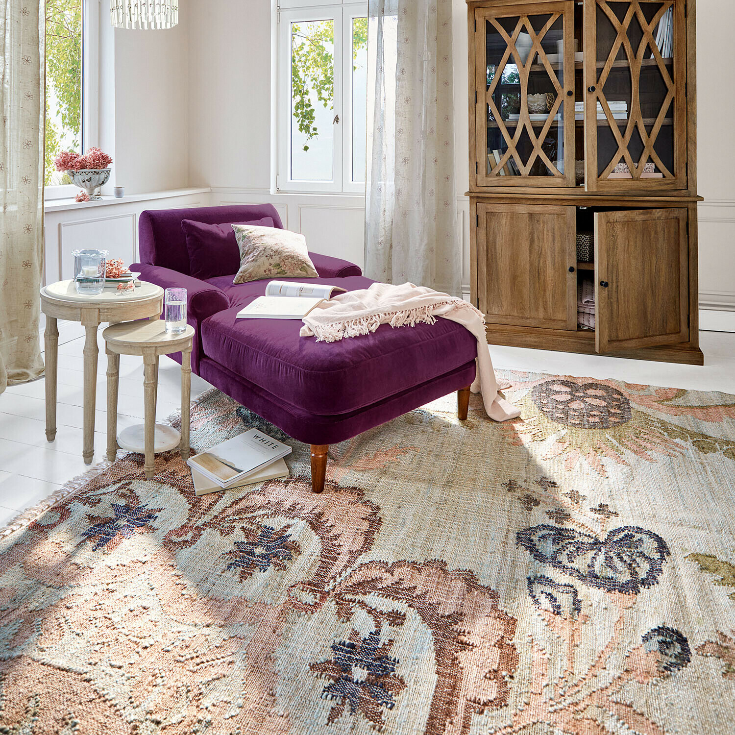 Teppich Amélorie Violette Samt-Chaiselongue in hellem Wohnzimmer mit Holzschrank und dekorativem Teppich