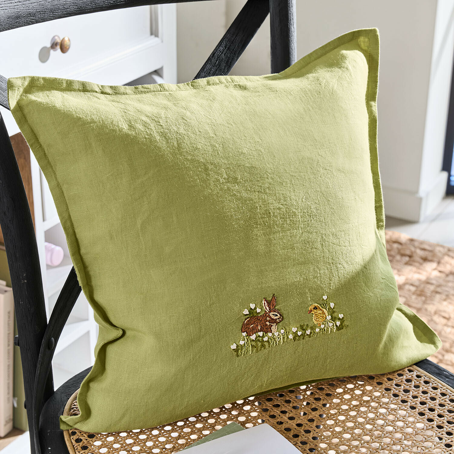 Grünes Leinenkissen mit Stickerei von Hase und Küken auf geflochtenem Stuhl, Wohnaccessoire