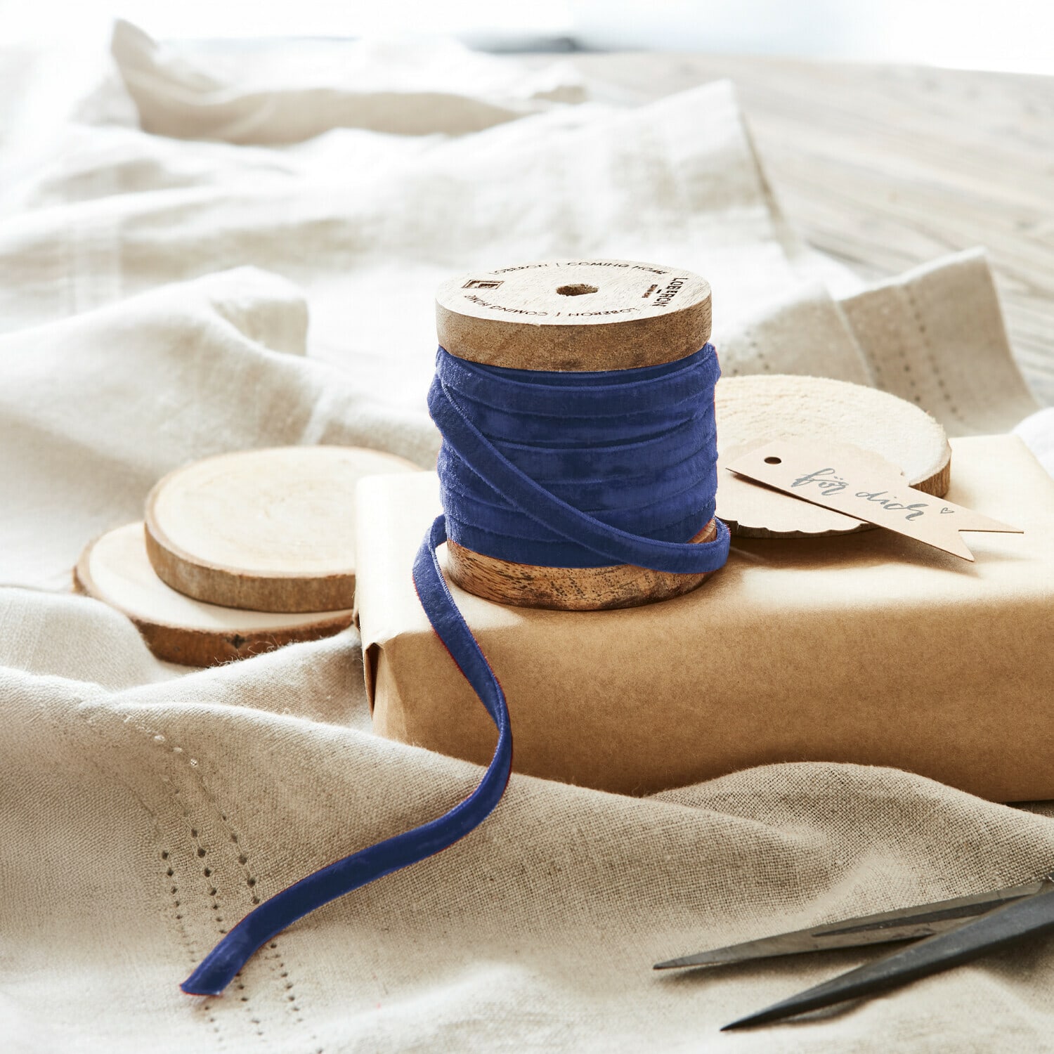 Samtband Olessa Blaue Samtschleife auf Holzspule vor Naturpapier und Stoff zum Geschenkverpacken