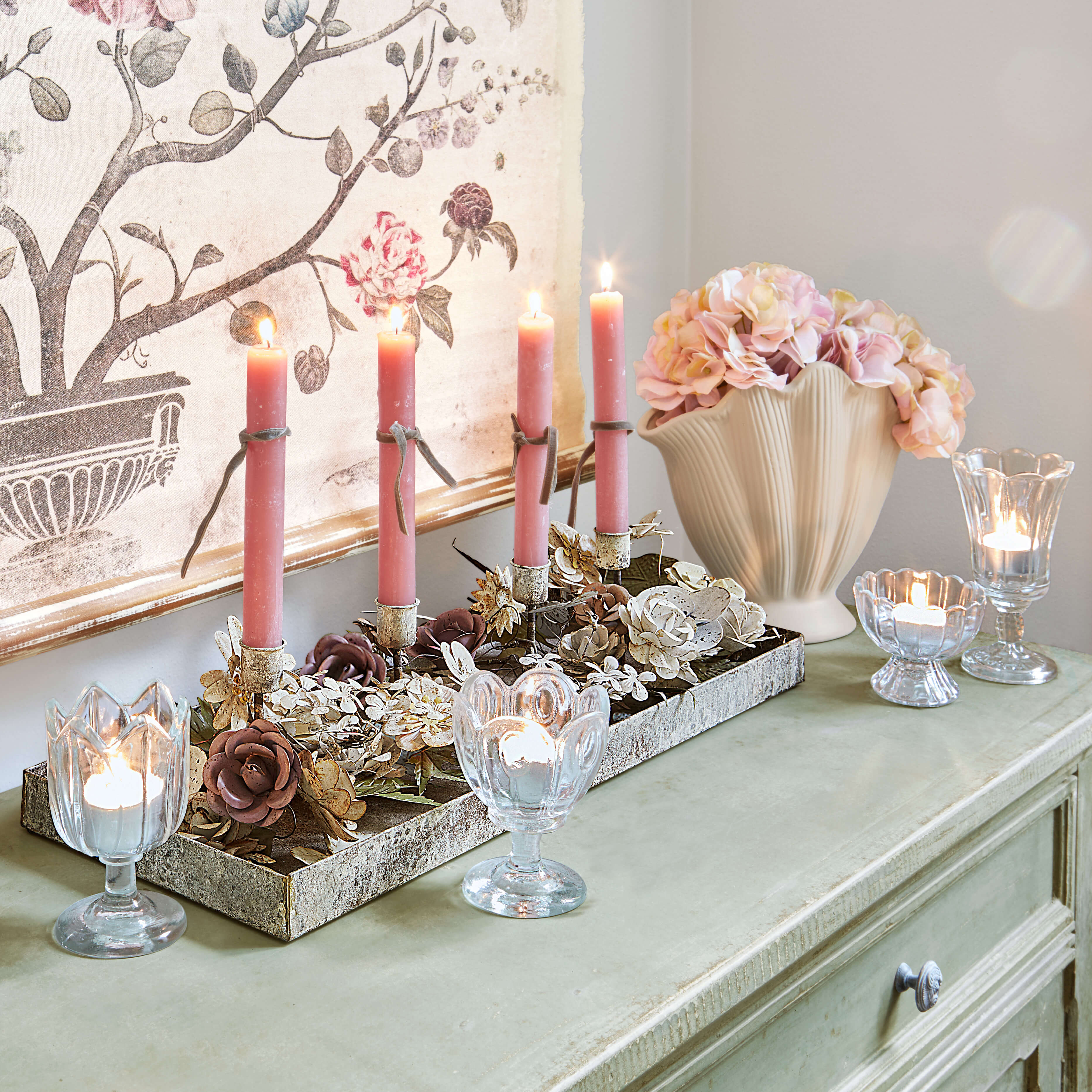 Dekorativer Kerzenhalter mit rosa Kerzen und Glaswindlichtern auf grünem Sideboard mit Blumen
