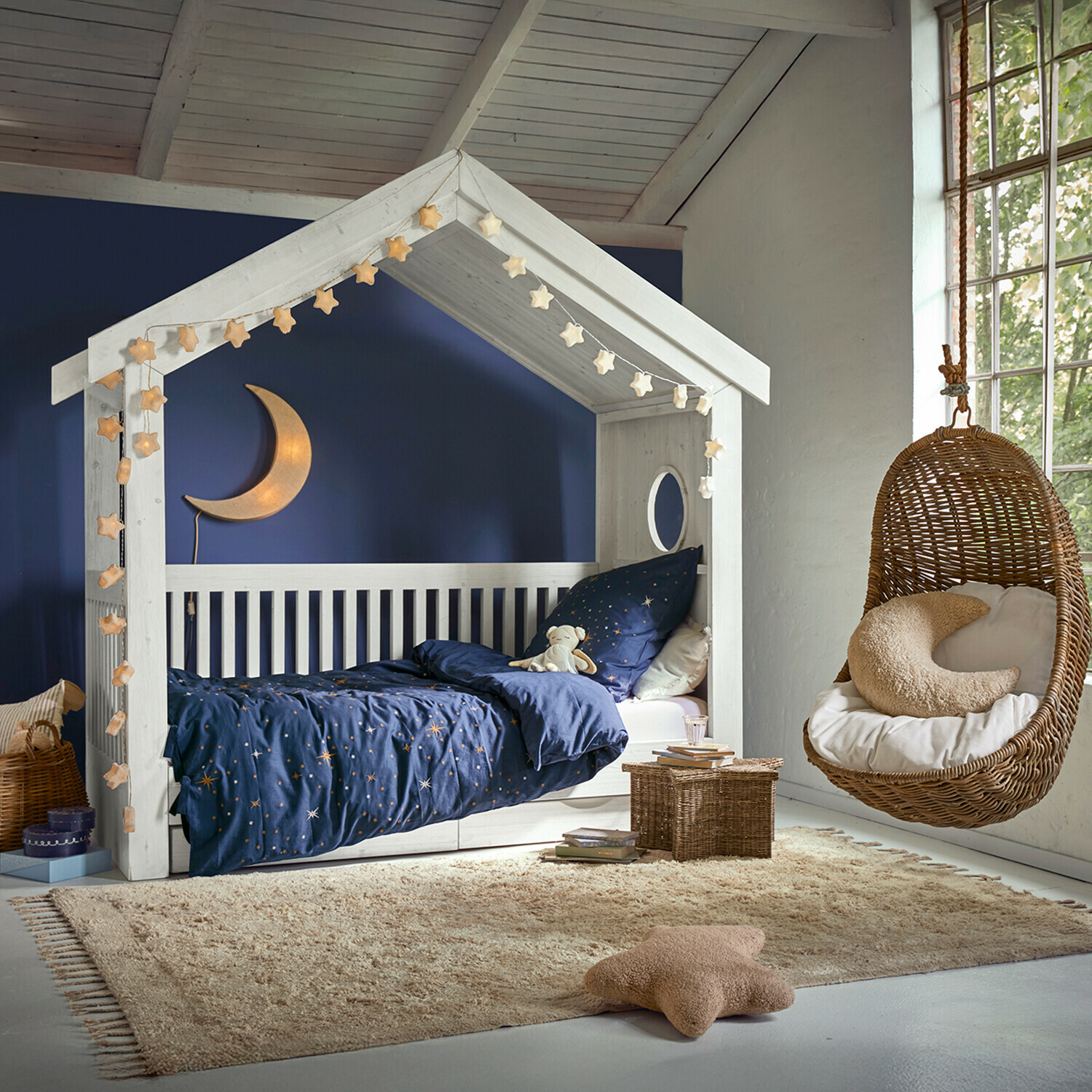 Weißes Holzhaus-Bett mit dunkelblauer Sternenbettwäsche und hängendem Korbsessel im Kinderzimmer