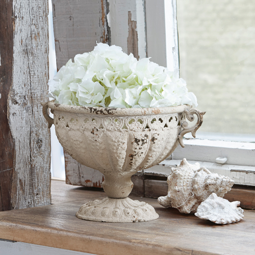 Dekorative Amphore Vescemont aus cremefarbenem Metall mit weißen Blumen auf Holzfensterbank