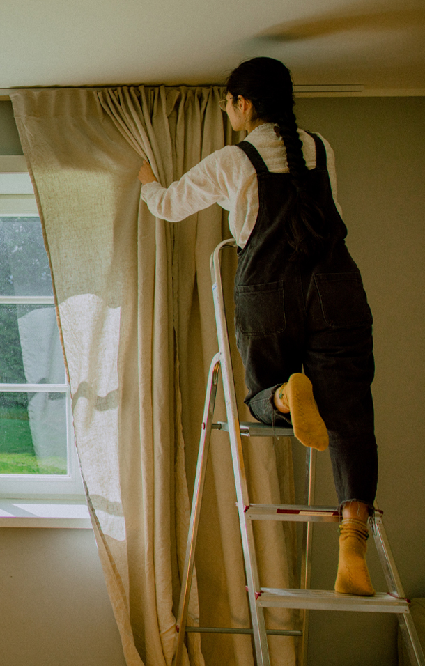 Frau auf Leiter zieht beigen Leinenvorhang vor Fenster in hellem Raum zu