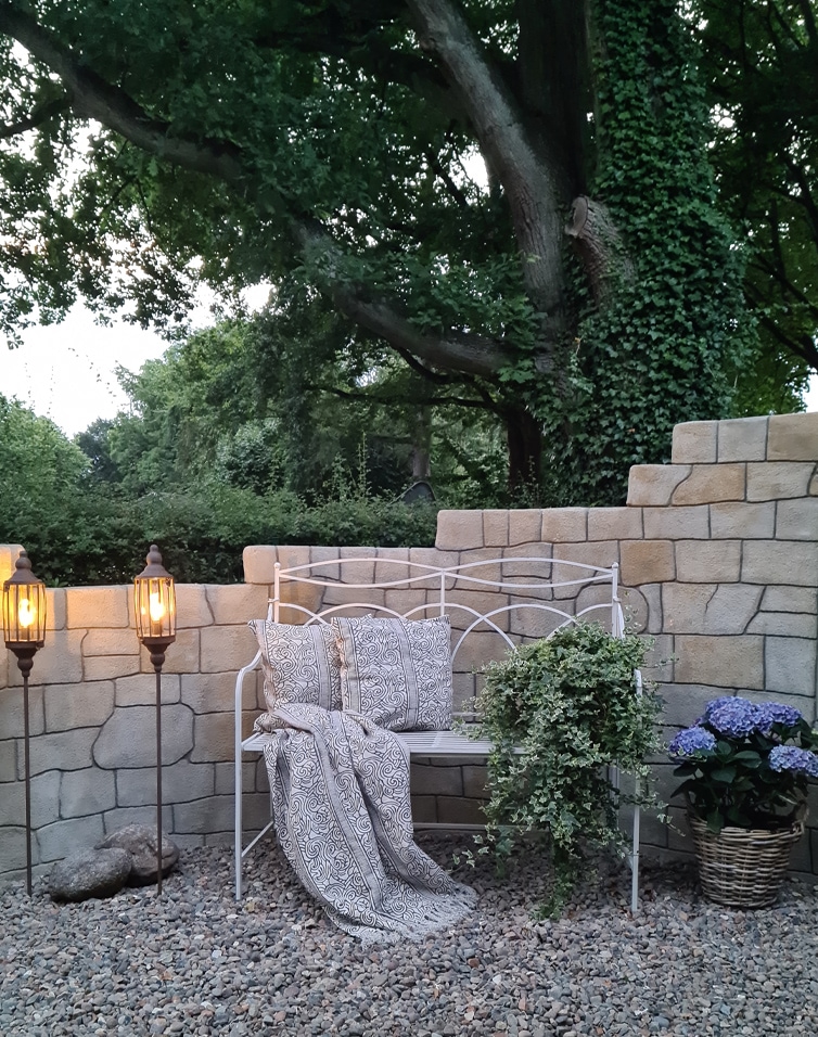 Weiße Metall-Gartenbank mit Kissen und Decke vor Steinmauer, umgeben von Pflanzen und Laternen