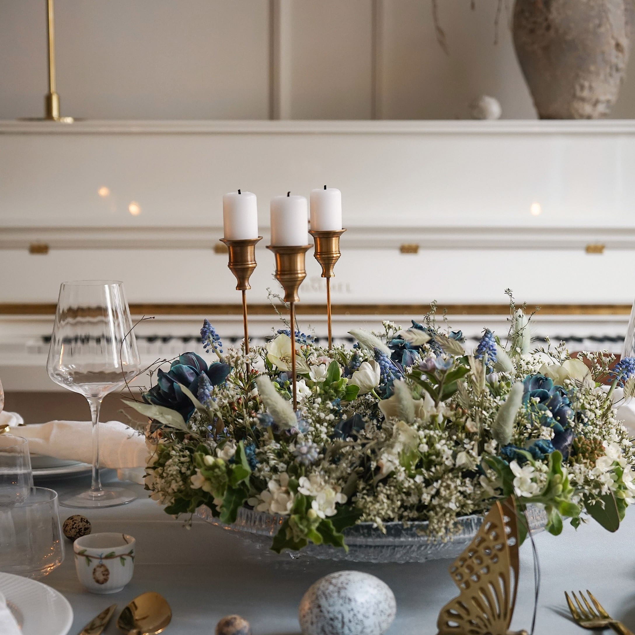 Dekorativer Kerzenhalter mit drei weißen Kerzen und Blumenarrangement auf Glasplatte