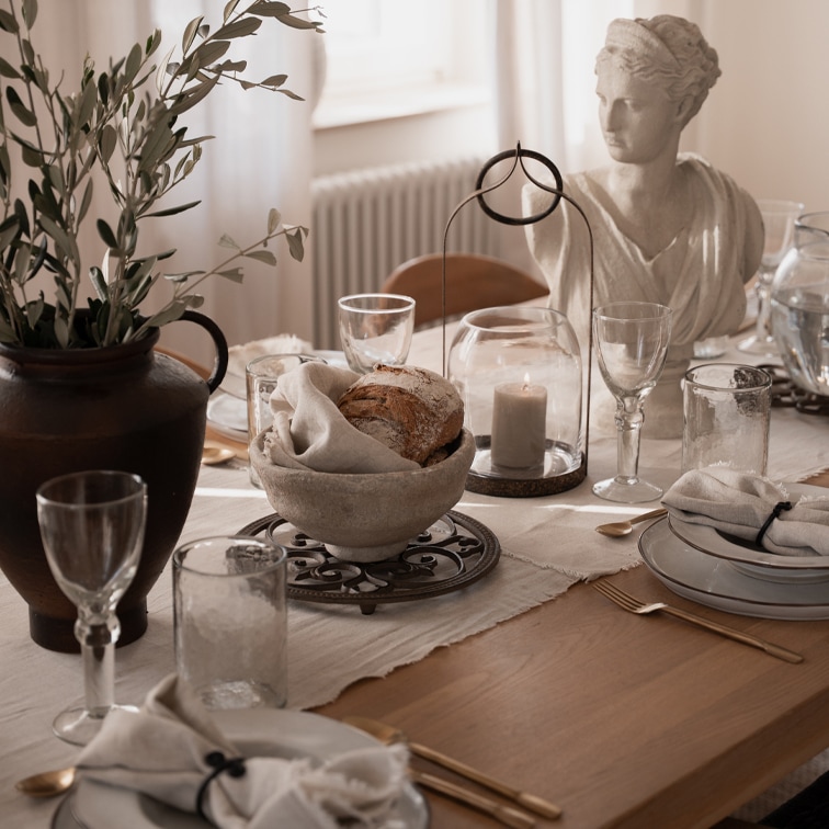 Rustikaler Tisch mit steinerner Brotschale, Kerze unter Glasglocke und weißer Steinskulptur als Dekor