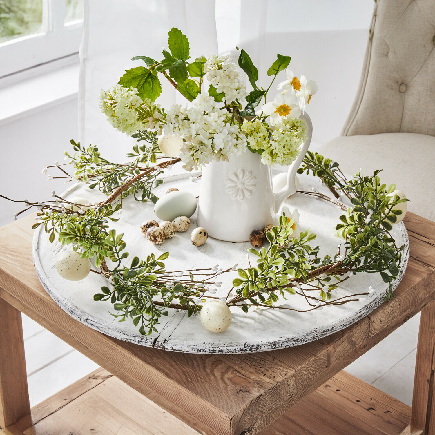 Dekorativer Kranz aus grünen Blättern und Eiern auf weißem Holztablett mit Keramikkrug und Blumen