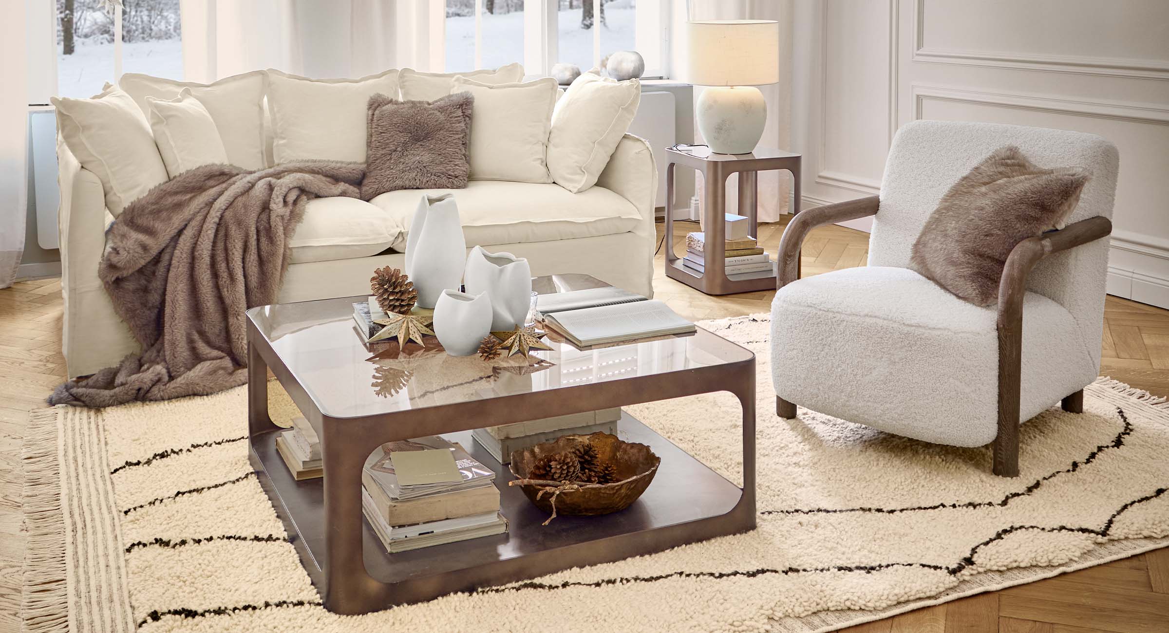 Gemütliches Wohnzimmer mit weißem Sofa, Sessel und braunem Kuscheldecke auf gemustertem Teppich