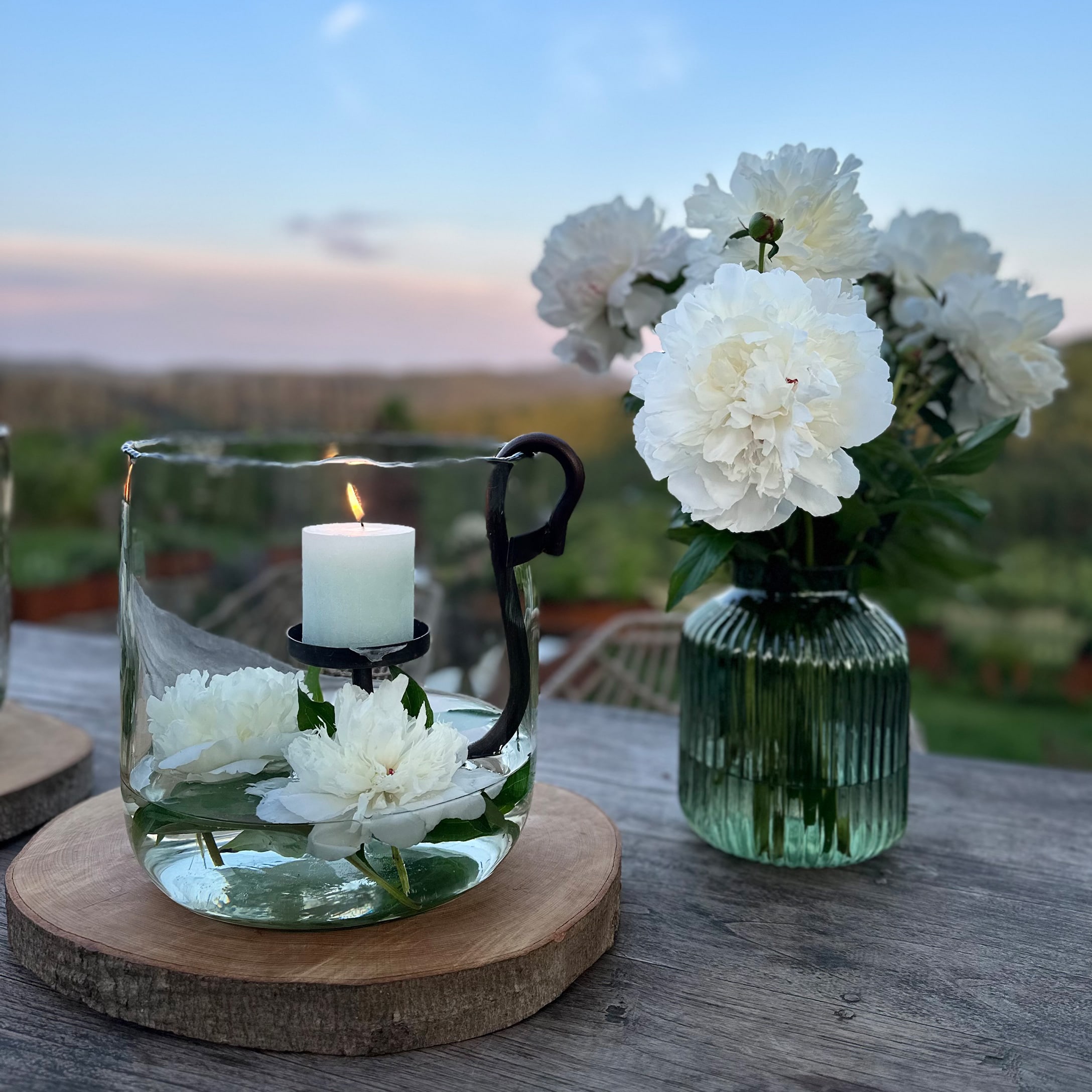 Glaswindlicht mit weißer Kerze und weißen Blumen auf Holzunterlage im Außenbereich