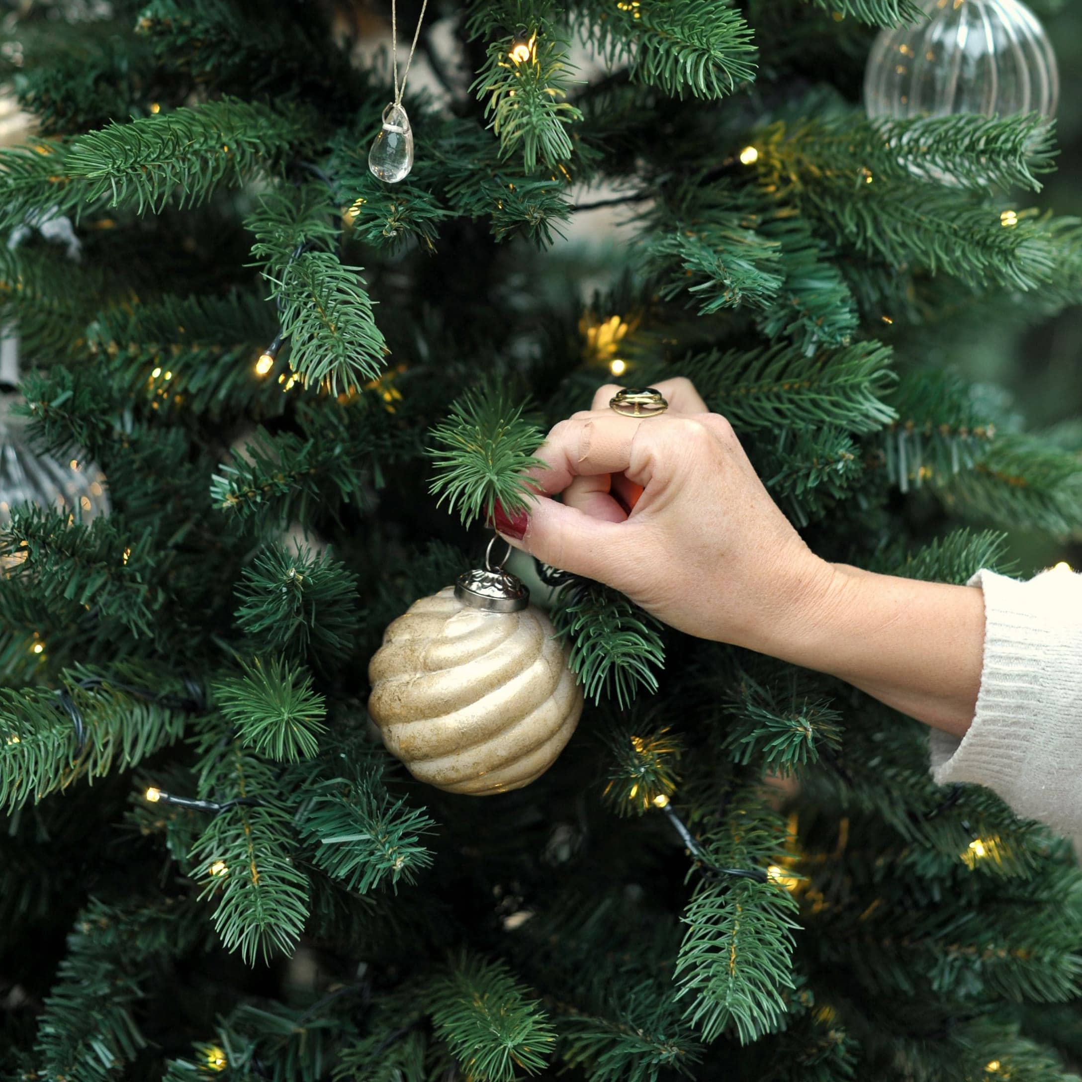 Hand schmückt grünen Weihnachtsbaum mit goldener Spiral-Kugel und LED-Lichterkette im Wintergarten