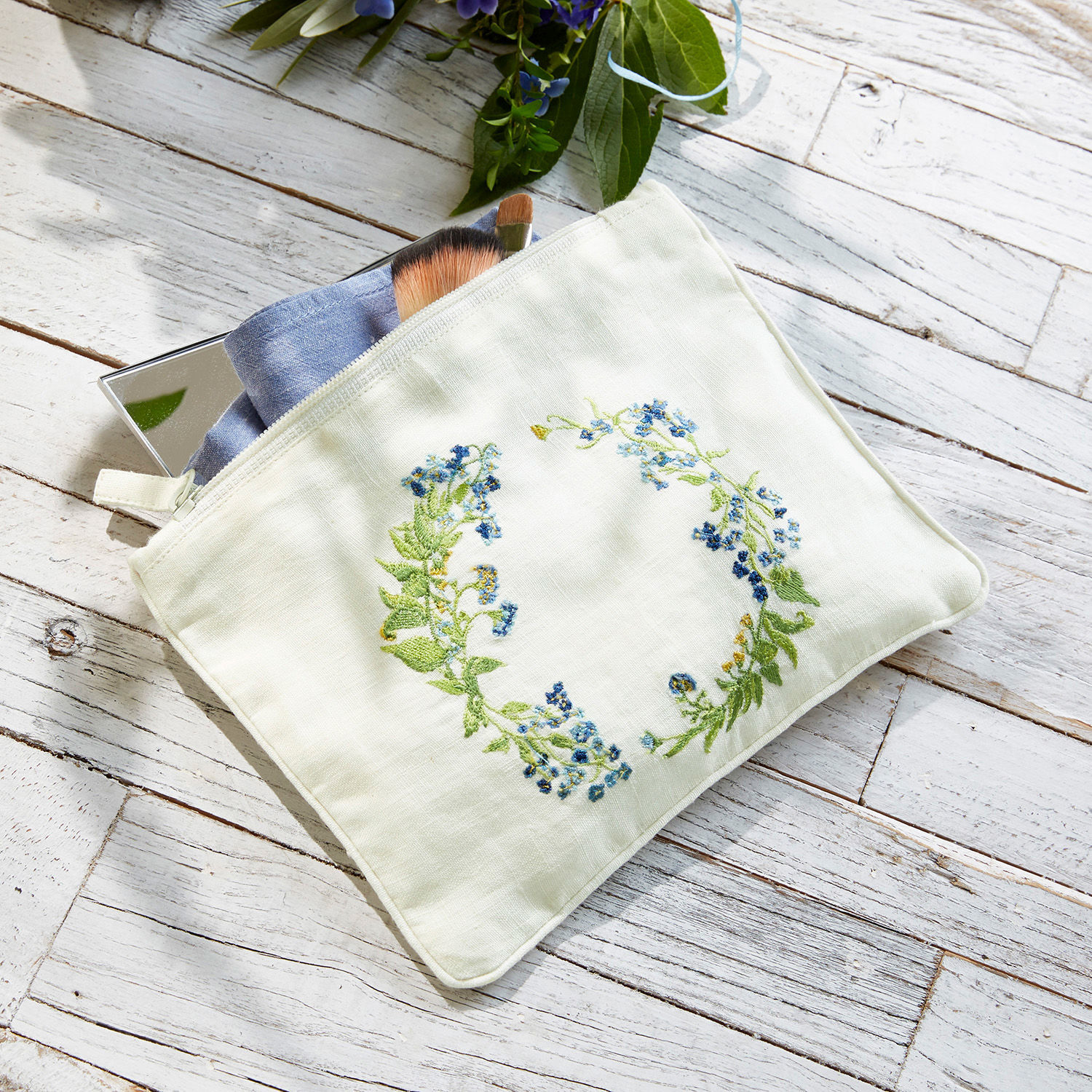 Täschchen Fierolla Weiße Kosmetiktasche aus Leinen mit blauer Blumenstickerei auf hellem Holzhintergrund