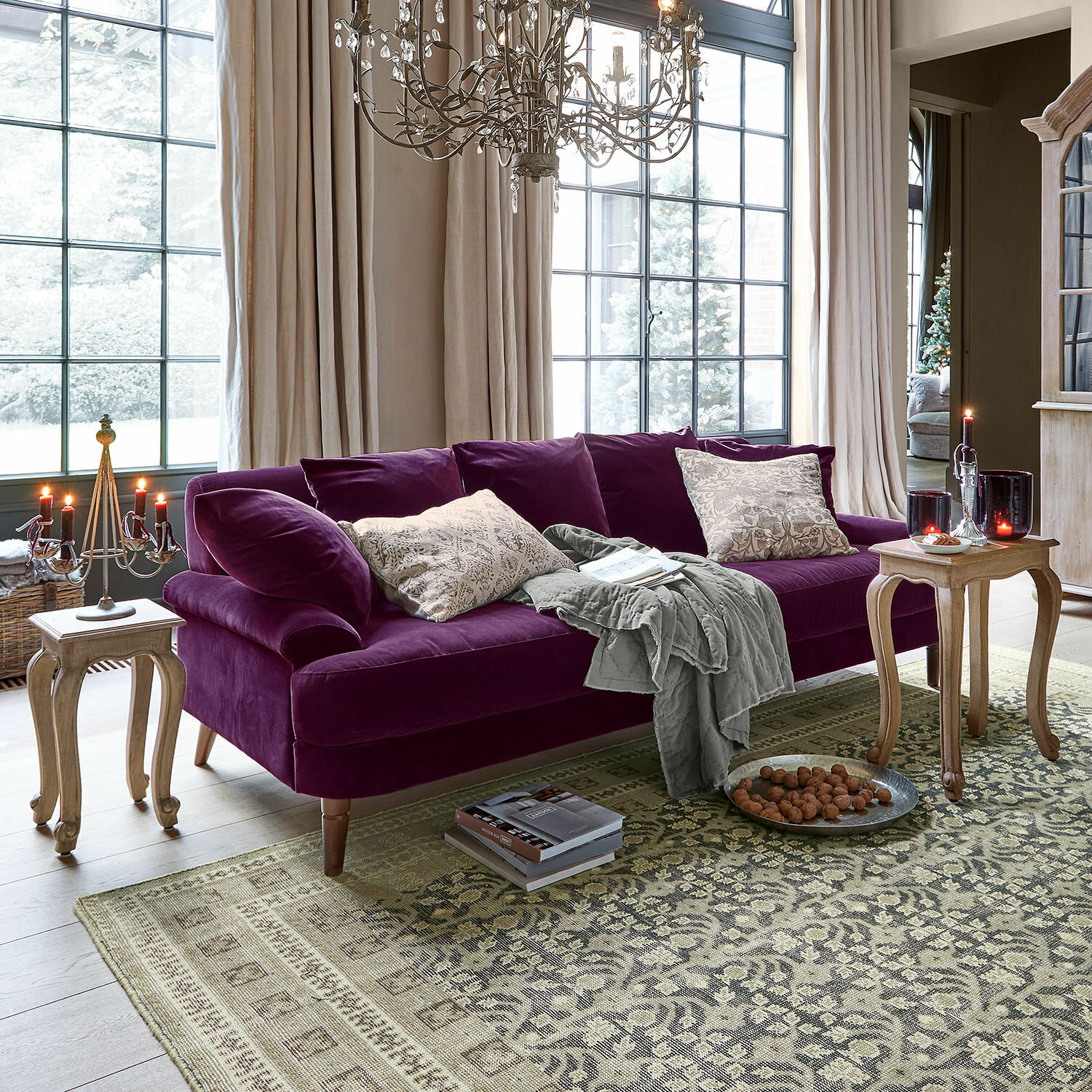 Sofa Rivermere Violette Samtsofa mit Kissen und grauer Decke in hellem Wohnzimmer mit großen Fenstern und Holztischen