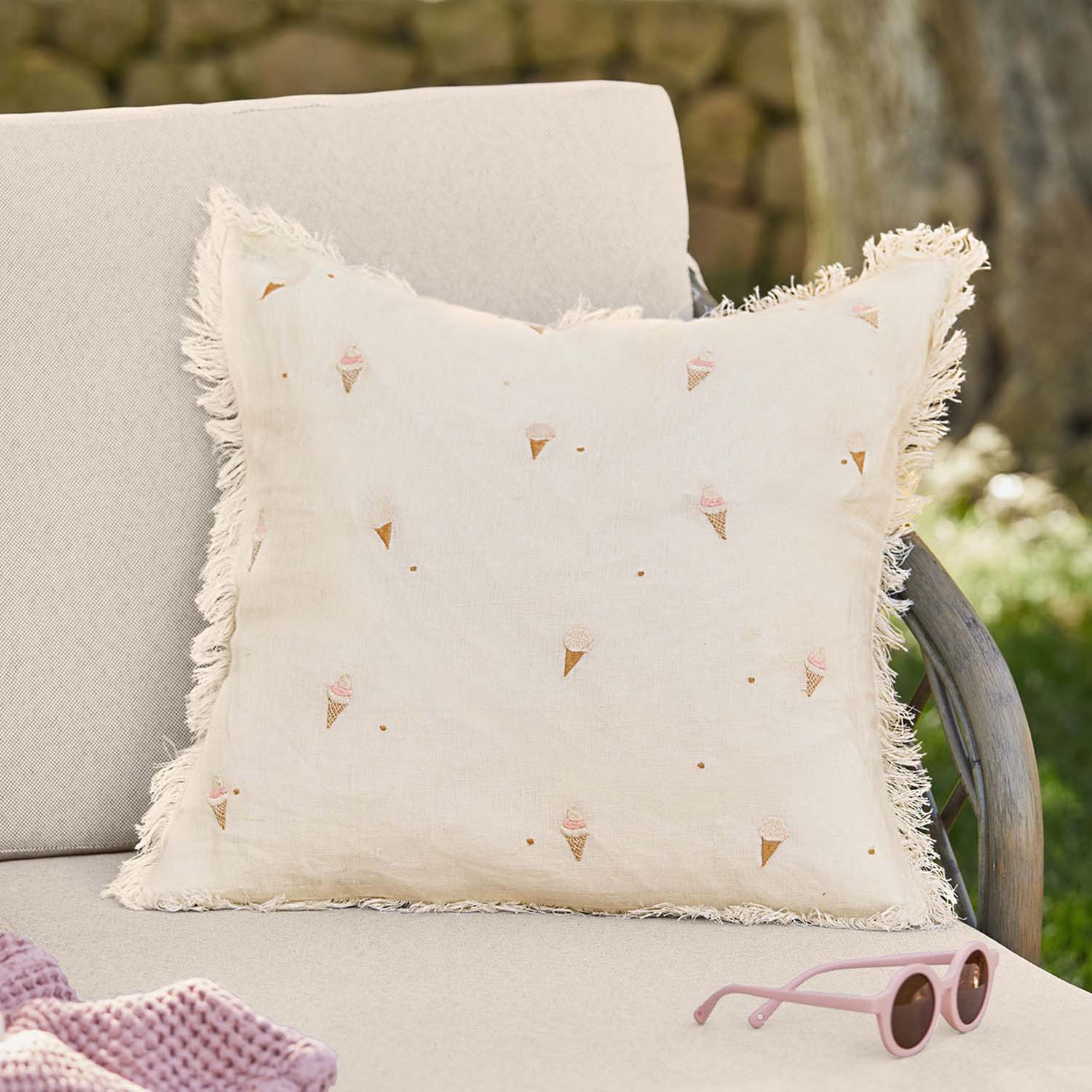 Beige Leinenkissen mit Fransen und pastellfarbenen Eistüten-Motiven auf Gartenbank