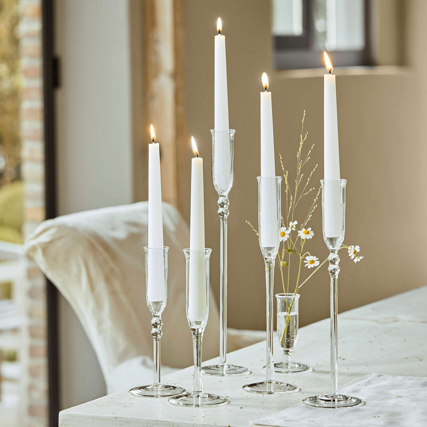 Sechs transparente Glas-Kerzenhalter mit weißen Kerzen auf hellem Tisch arrangiert