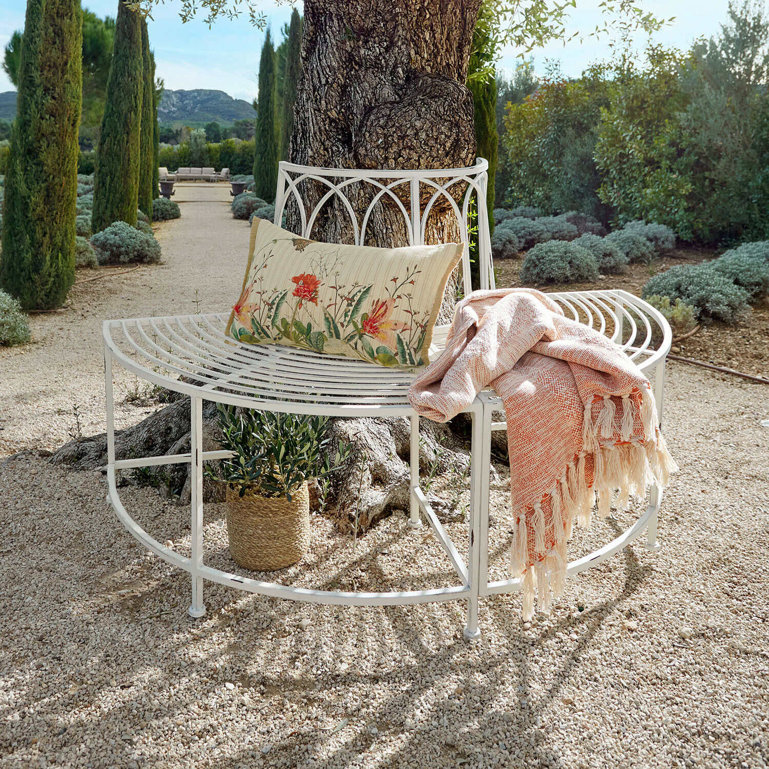 Weiße Metallbank um einen Baum mit floralem Kissen und orangefarbenem Plaid auf Kiesweg im Garten
