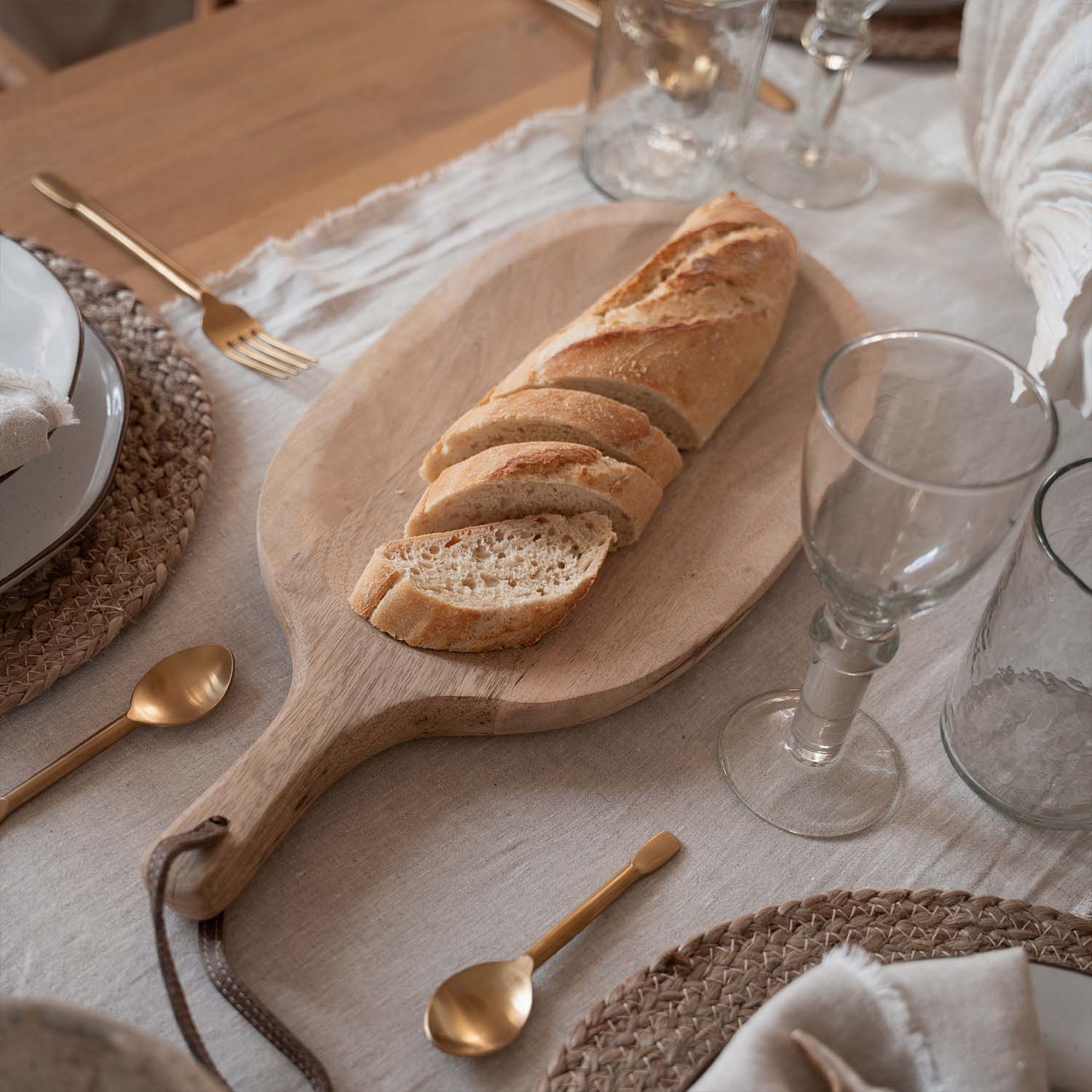 Holzschneidebrett mit aufgeschnittener Baguette auf Tisch mit natürlichem Leinentuch und goldenen Bestecken