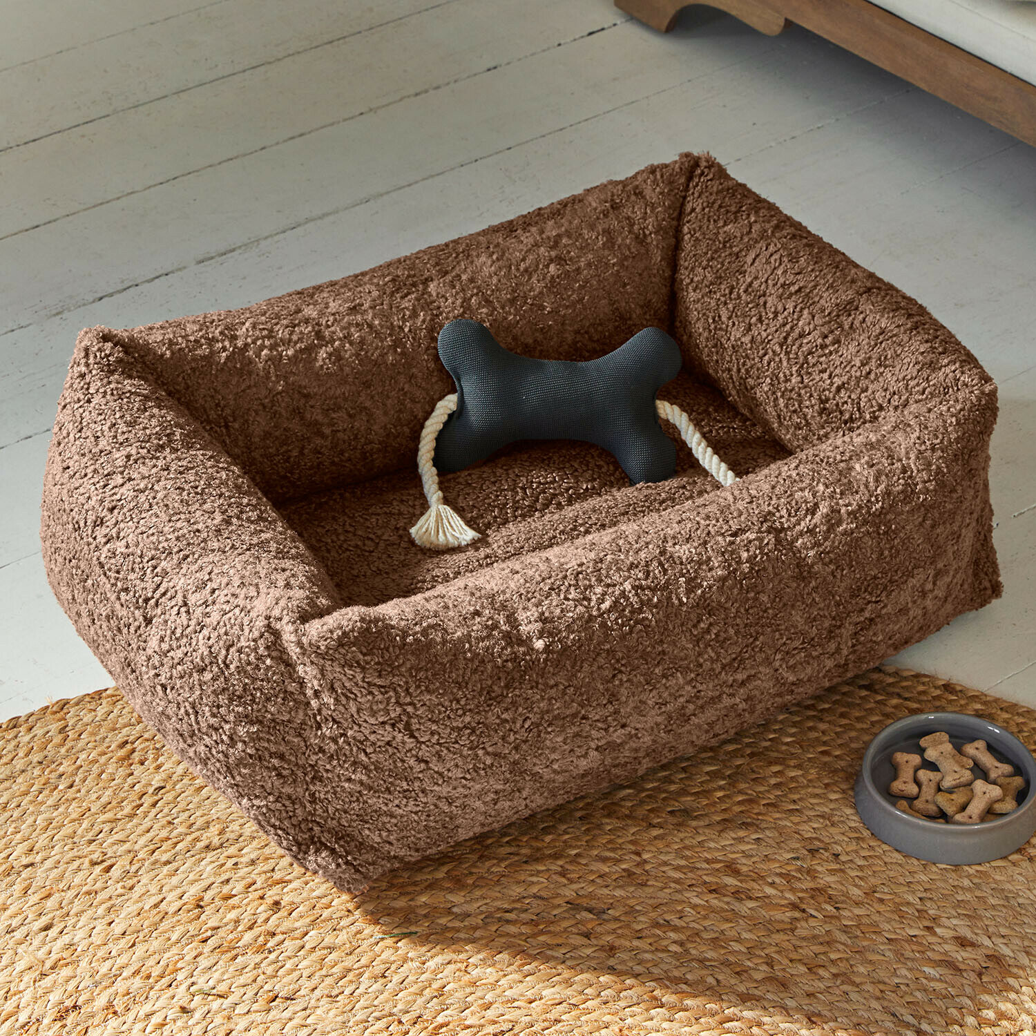Haustierbett Giolena Braunes Plüsch-Hundebett mit schwarzem Knochen-Spielzeug und Leckerli-Schale auf geflochtenem Teppich