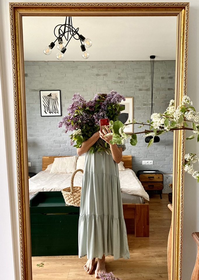 Frau im langen grünen Kleid hält lila Blumenstrauß vor Spiegel in Schlafzimmer mit Holzbett