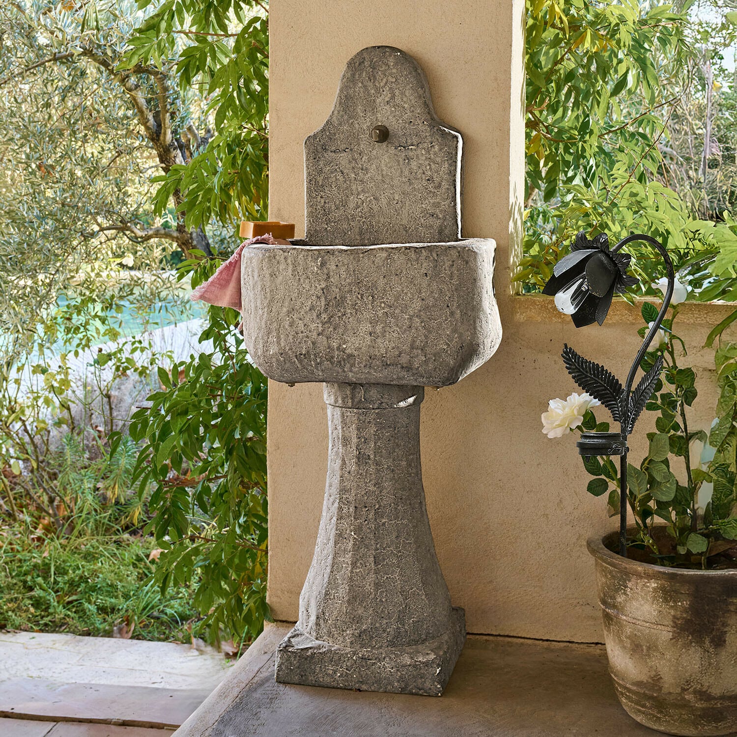 Brunnen Lurevino Dekorativer grauer Steingarten-Waschbecken auf Säule mit natürlicher Oberfläche und Pflanzen im Hintergrund
