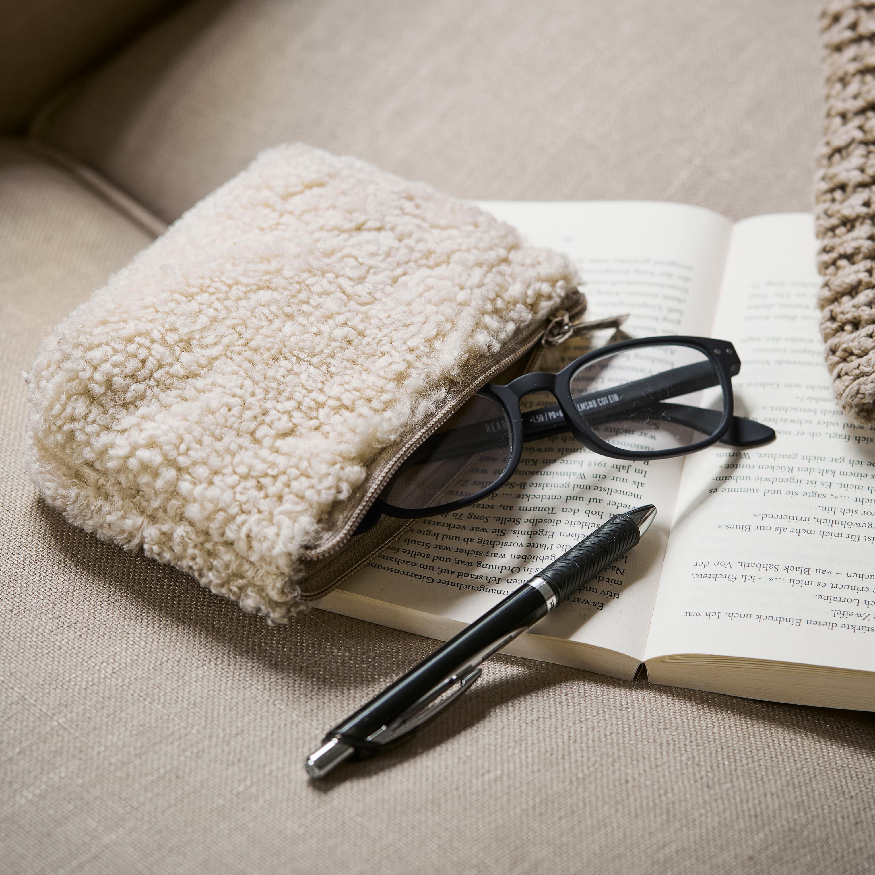 Beige, flauschige Kosmetiktasche auf Sofa neben Brille und Stift auf offenem Buch