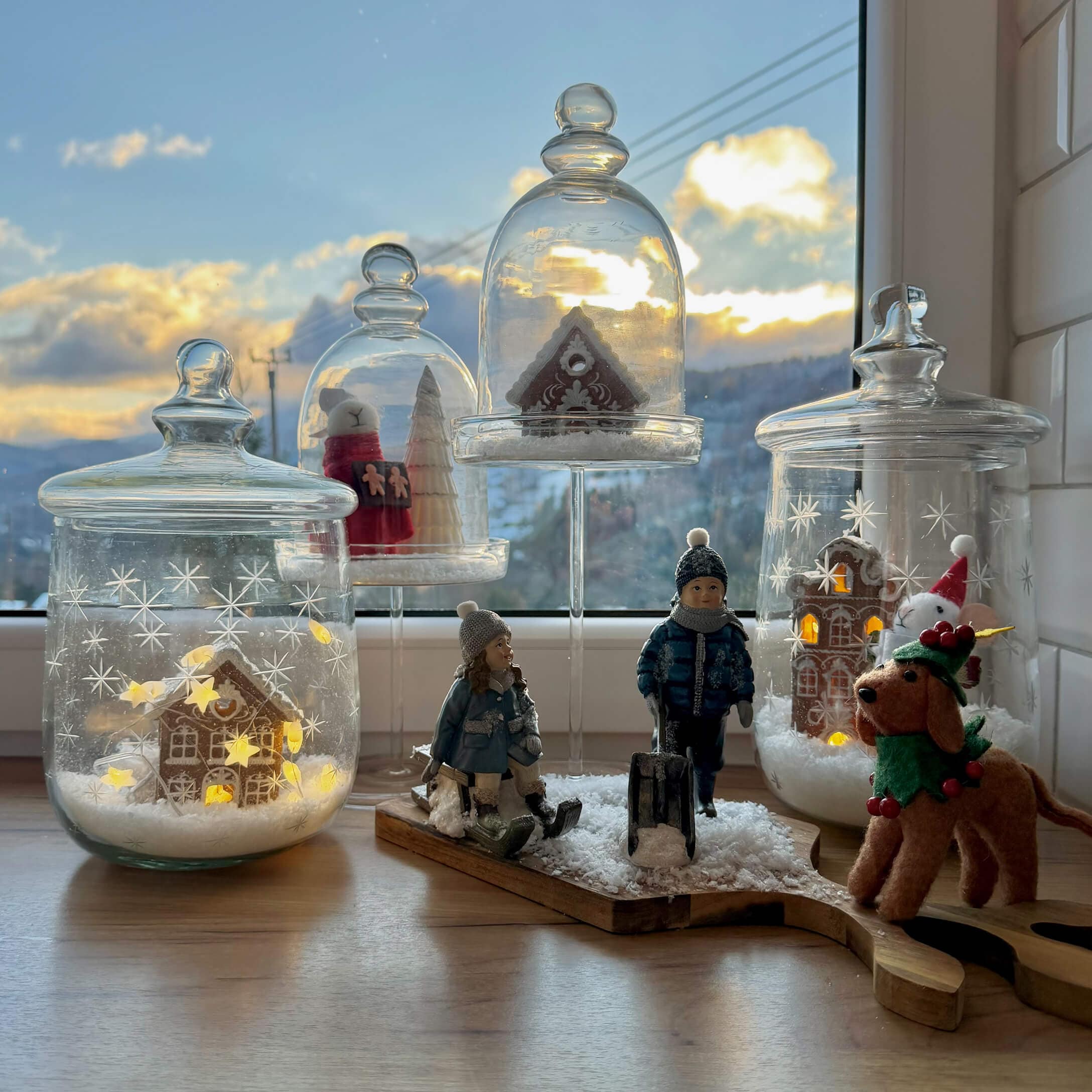 Weihnachtsdekoration mit Kinderfiguren, Hund und Lebkuchenhäusern unter Glaskuppeln auf Fensterbank