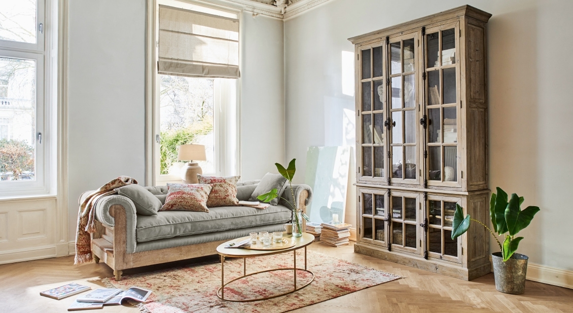 Helles Wohnzimmer mit grauem Sofa und großer Holzvitrine mit Glastüren, Teppich und Pflanzen