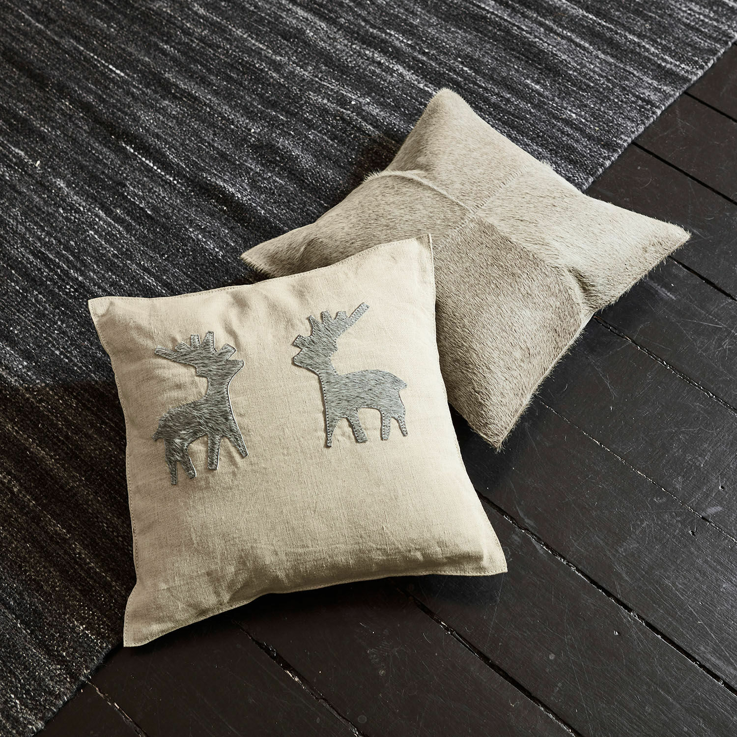 Kissenhülle 2er Set Beaupuy Beige Leinenkissen mit grauen Hirsch-Applikationen und hellbraunes Lederkissen auf dunklem Boden und Teppich