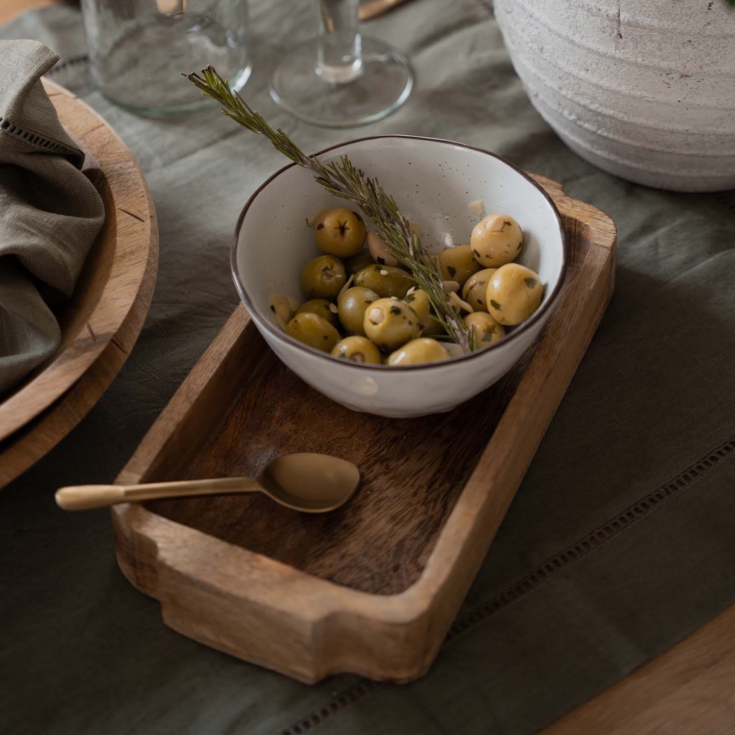 Weiße Keramikschale mit grünen Oliven und Rosmarinzweig auf Holztablett