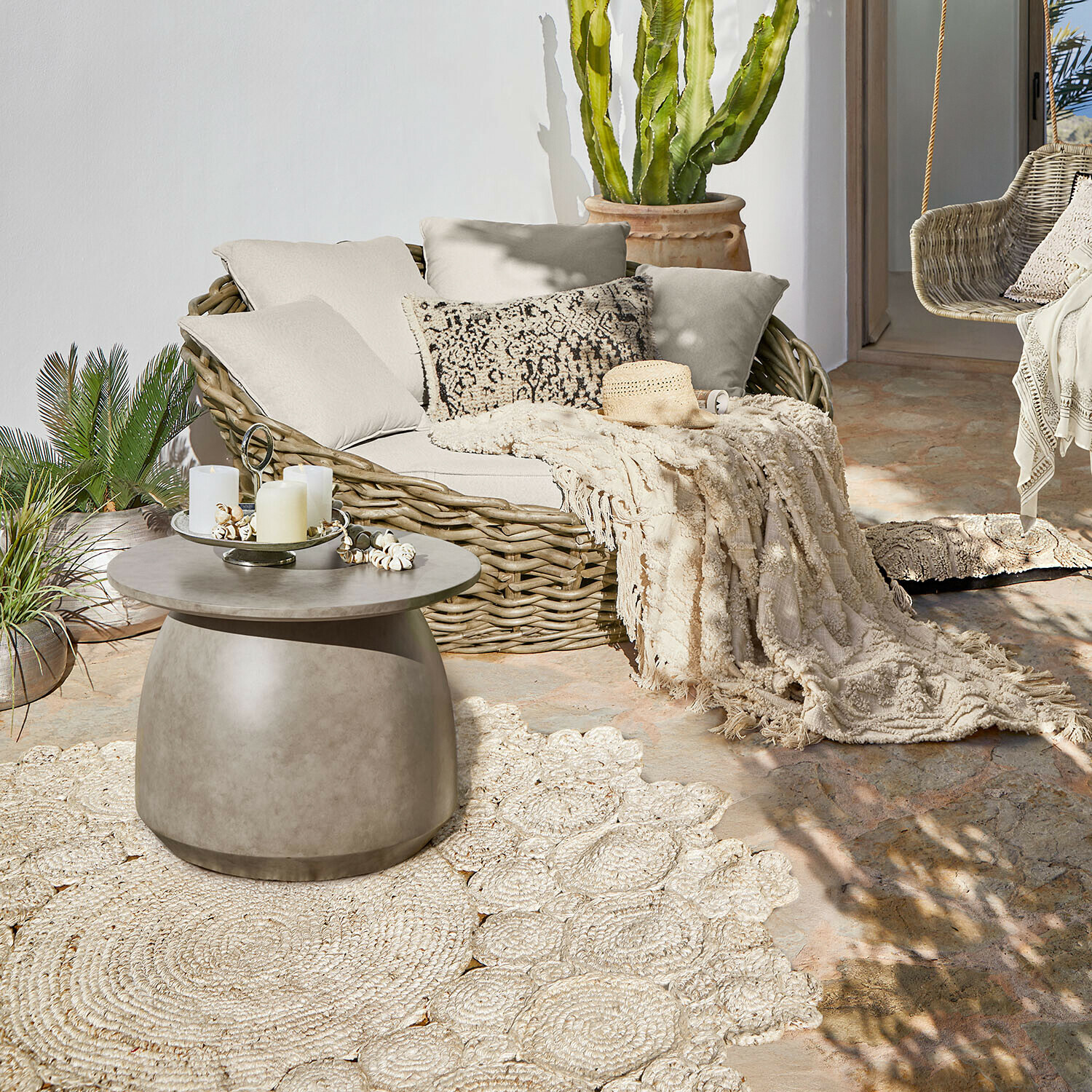 Ovales Rattansofa mit cremefarbenen Kissen und Decke auf Terrasse mit Beton-Couchtisch
