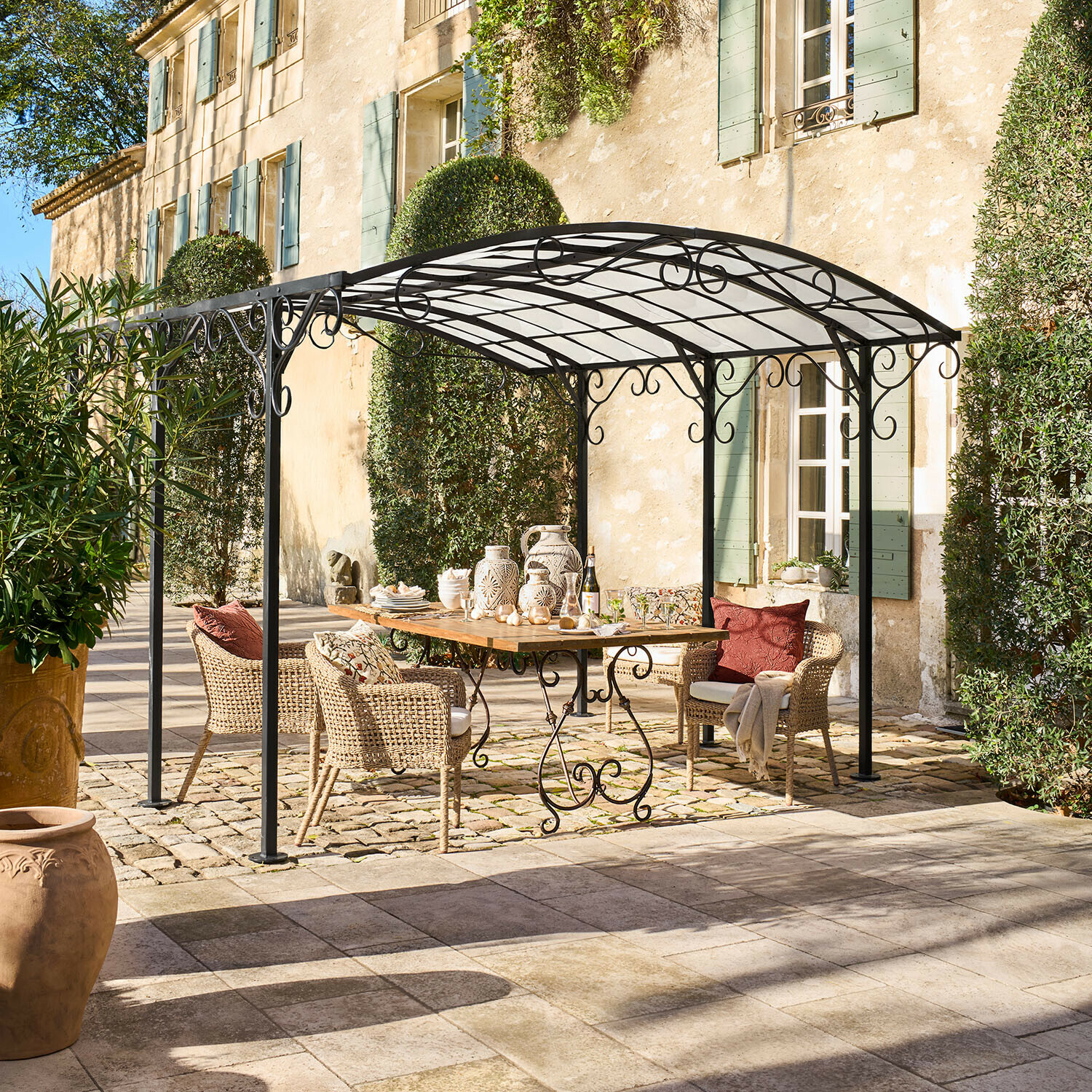 Pergola Delbridge Schwarze Metall-Gartenlaube mit dekorativen Schnörkeln über Holztisch und Rattanstühlen im Patio