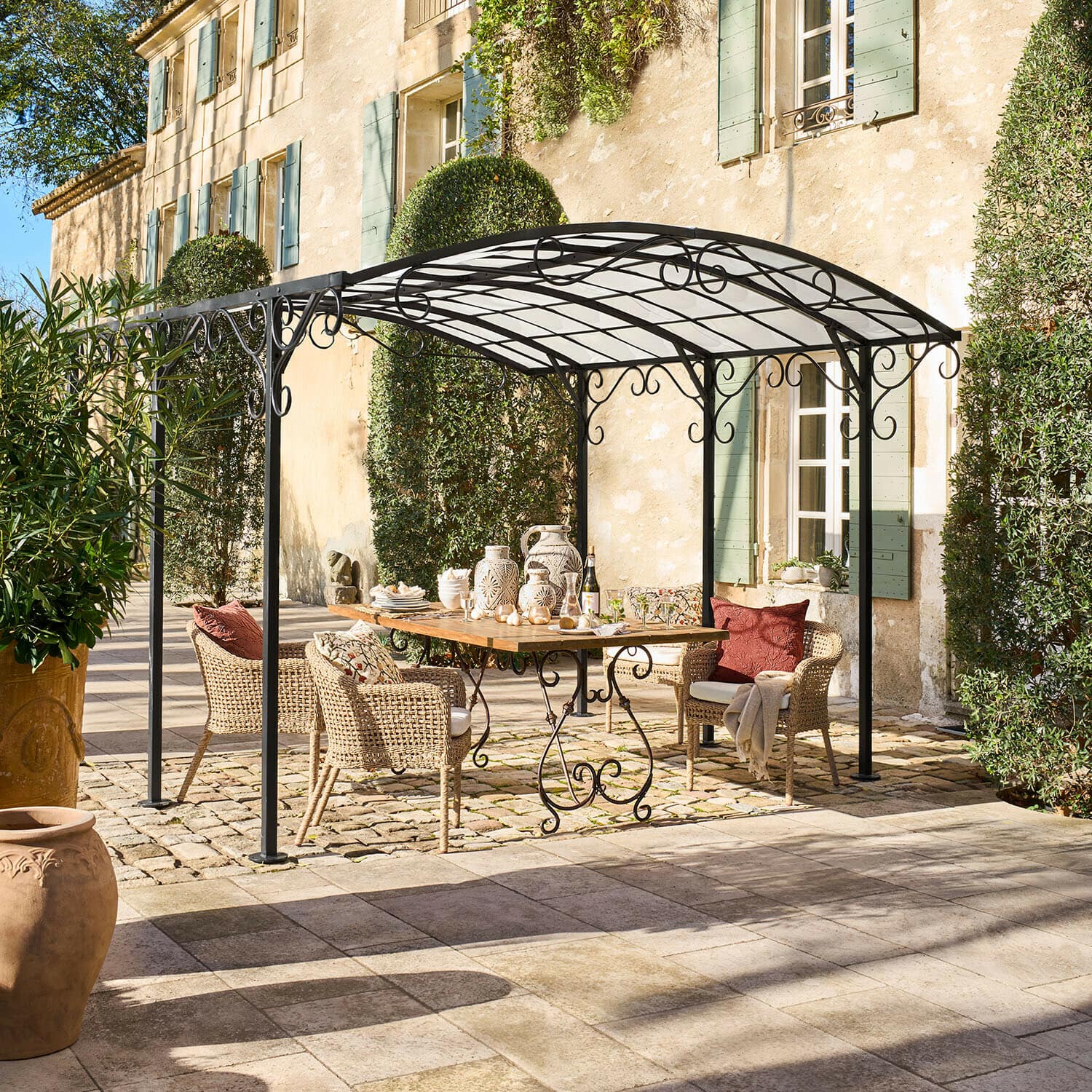 Pergola Delbridge Schwarze Metall-Gartenlaube mit dekorativen Schnörkeln über Holztisch und Rattanstühlen im Patio