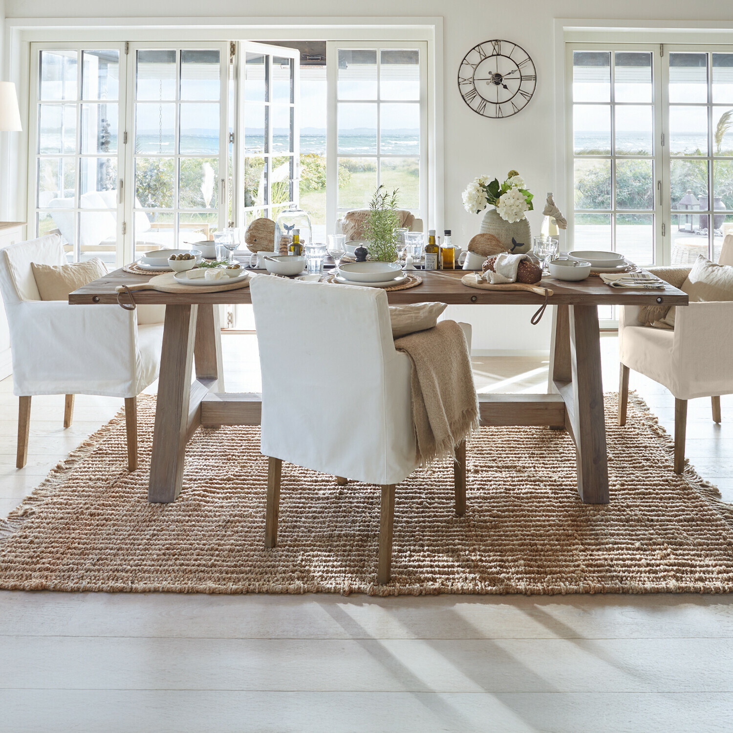 Esszimmer mit Holztisch und weißen Stühlen auf Naturteppich mit Blick auf Garten und Meer