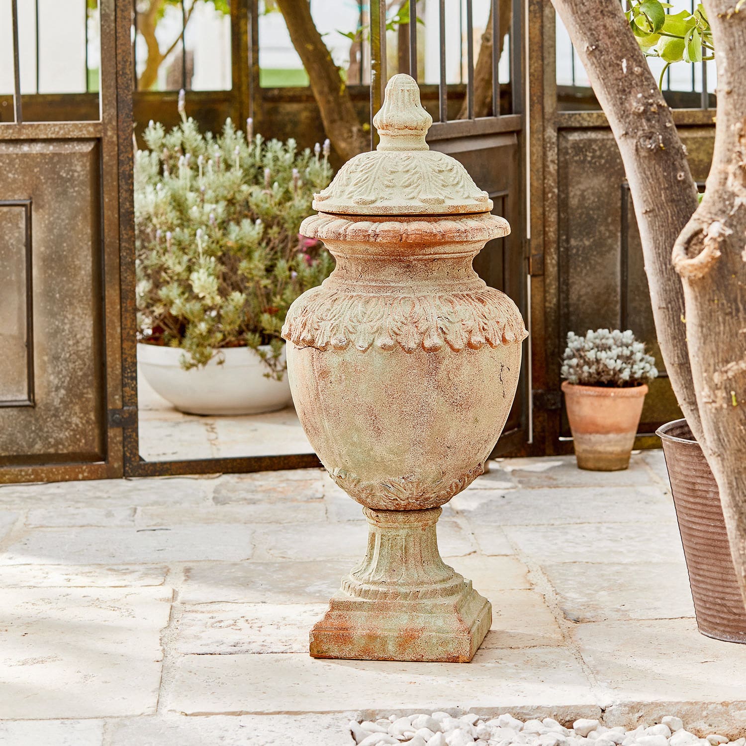 Deko-Objekt Persephone Dekorativer Garten-Vase aus Terrakotta in Naturfarbe auf Steinboden mit Pflanzen im Hintergrund