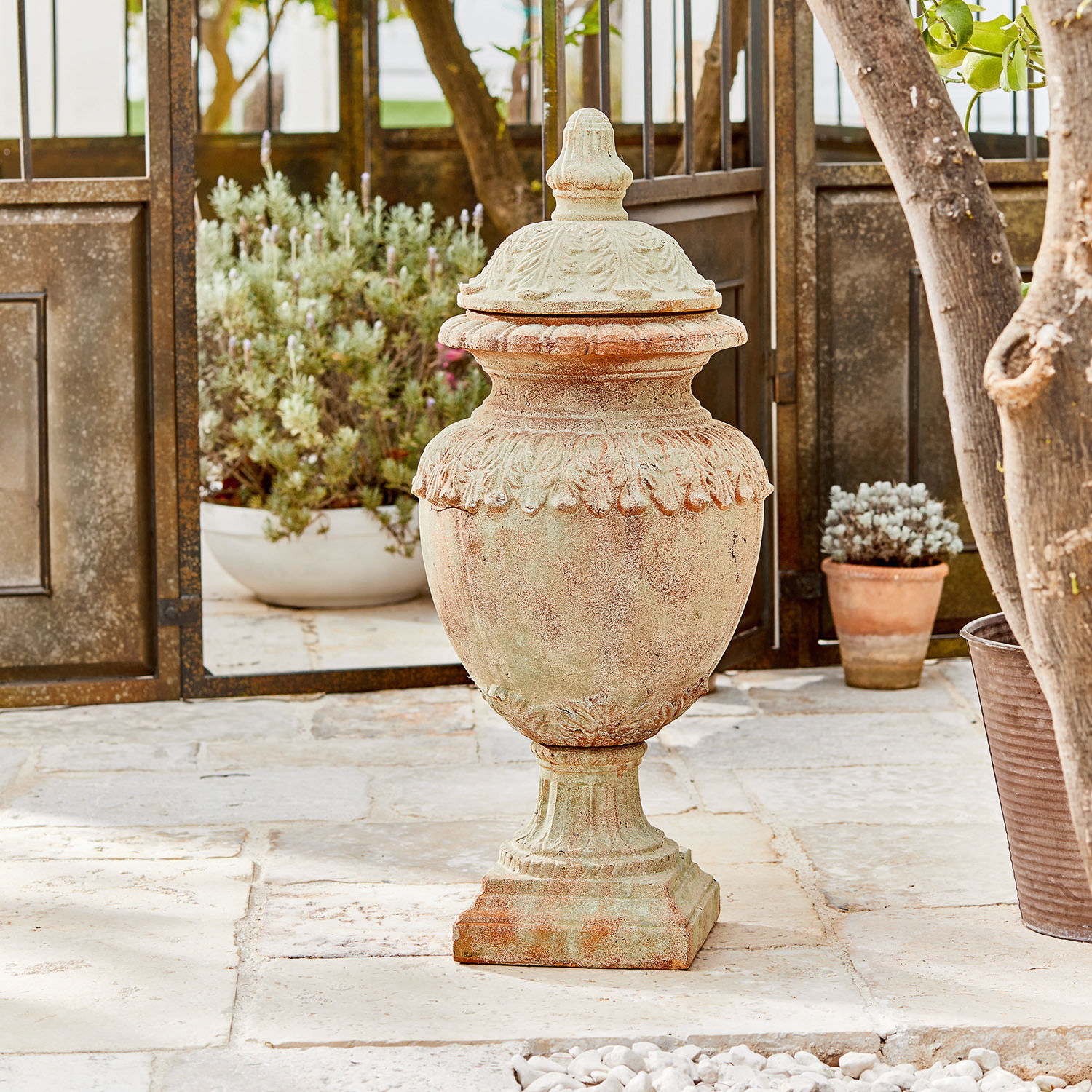 Deko-Objekt Persephone Dekorativer Garten-Vase aus Terrakotta in Naturfarbe auf Steinboden mit Pflanzen im Hintergrund